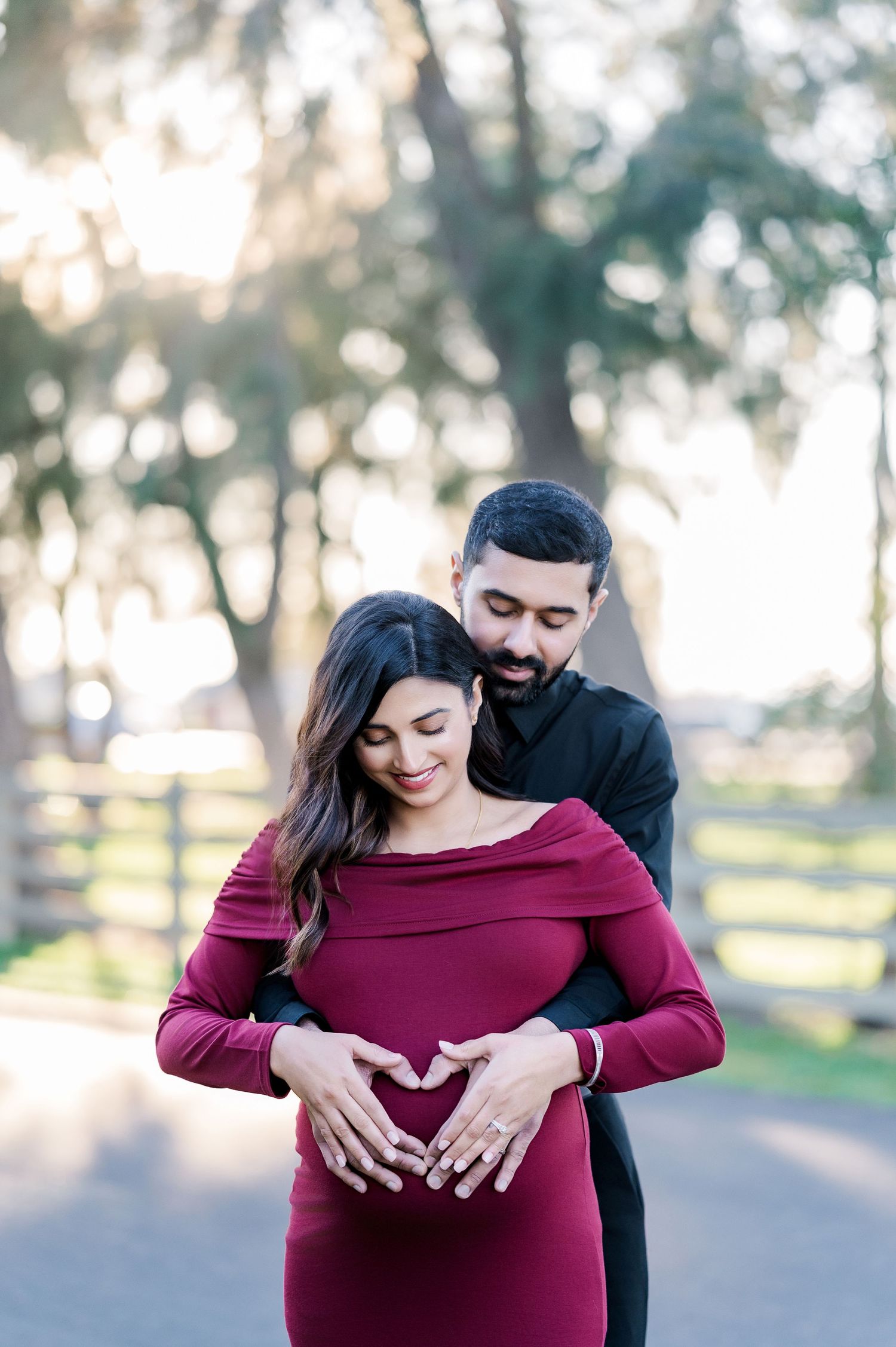 Maternity photoshoot in a park showing couple embracing with burgundy dress against sunlit trees and wooden fence.