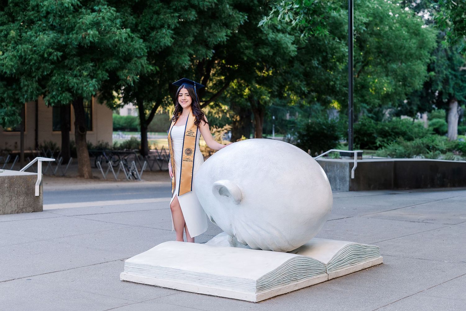 Graduate in regalia poses with a large decorative book sculpture on a university campus walkway.