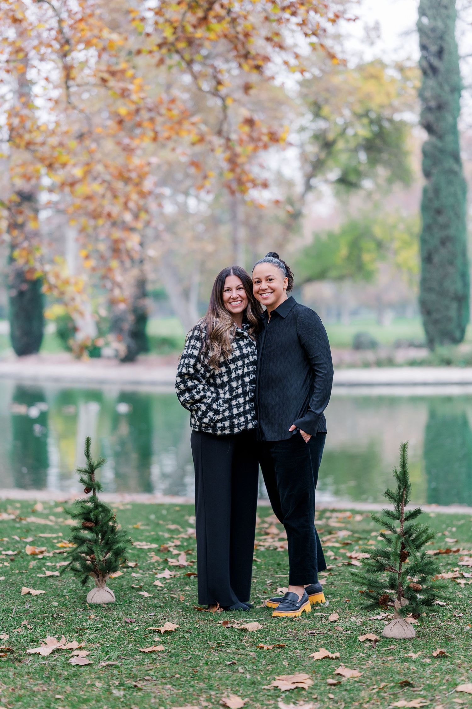Two people pose together in an autumn park setting with fallen leaves and a lake in the background.