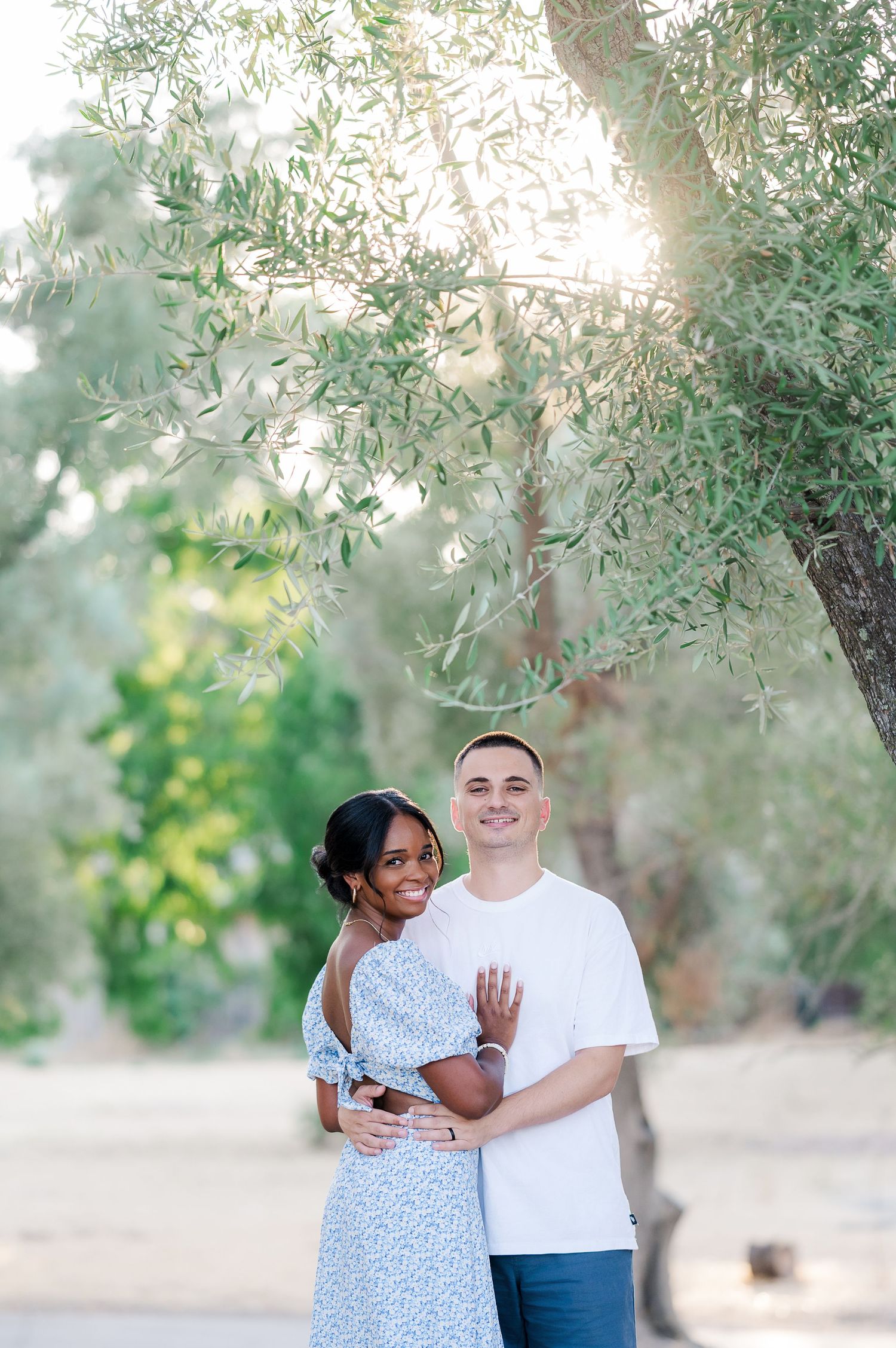 A couple shares a tender moment together while standing among olive trees in a sun-dappled outdoor setting.