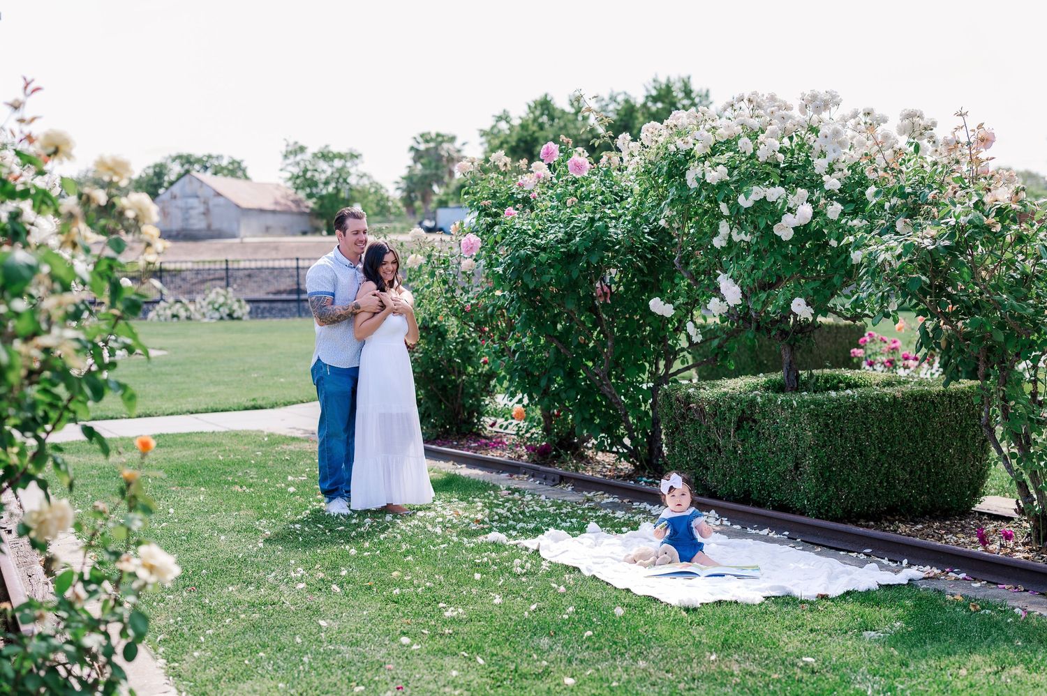 Family shares intimate moment in garden setting with white roses and hedges.