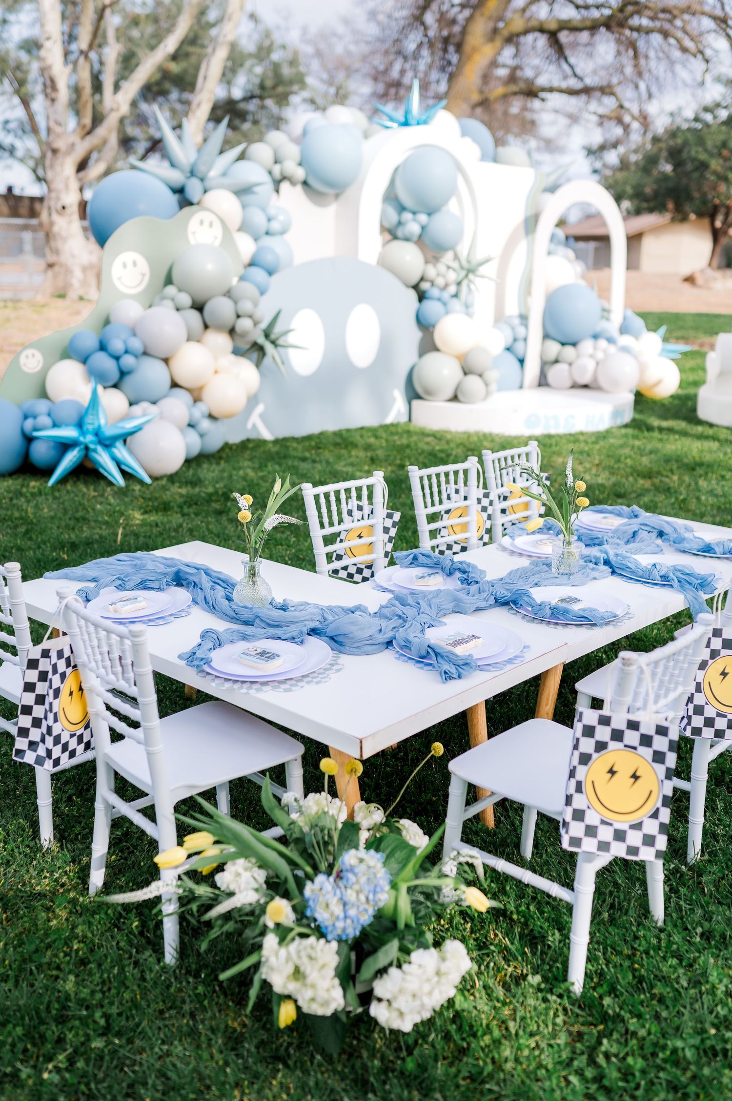 An elegant blue and white party table setting with balloon arch and smiley face decor.