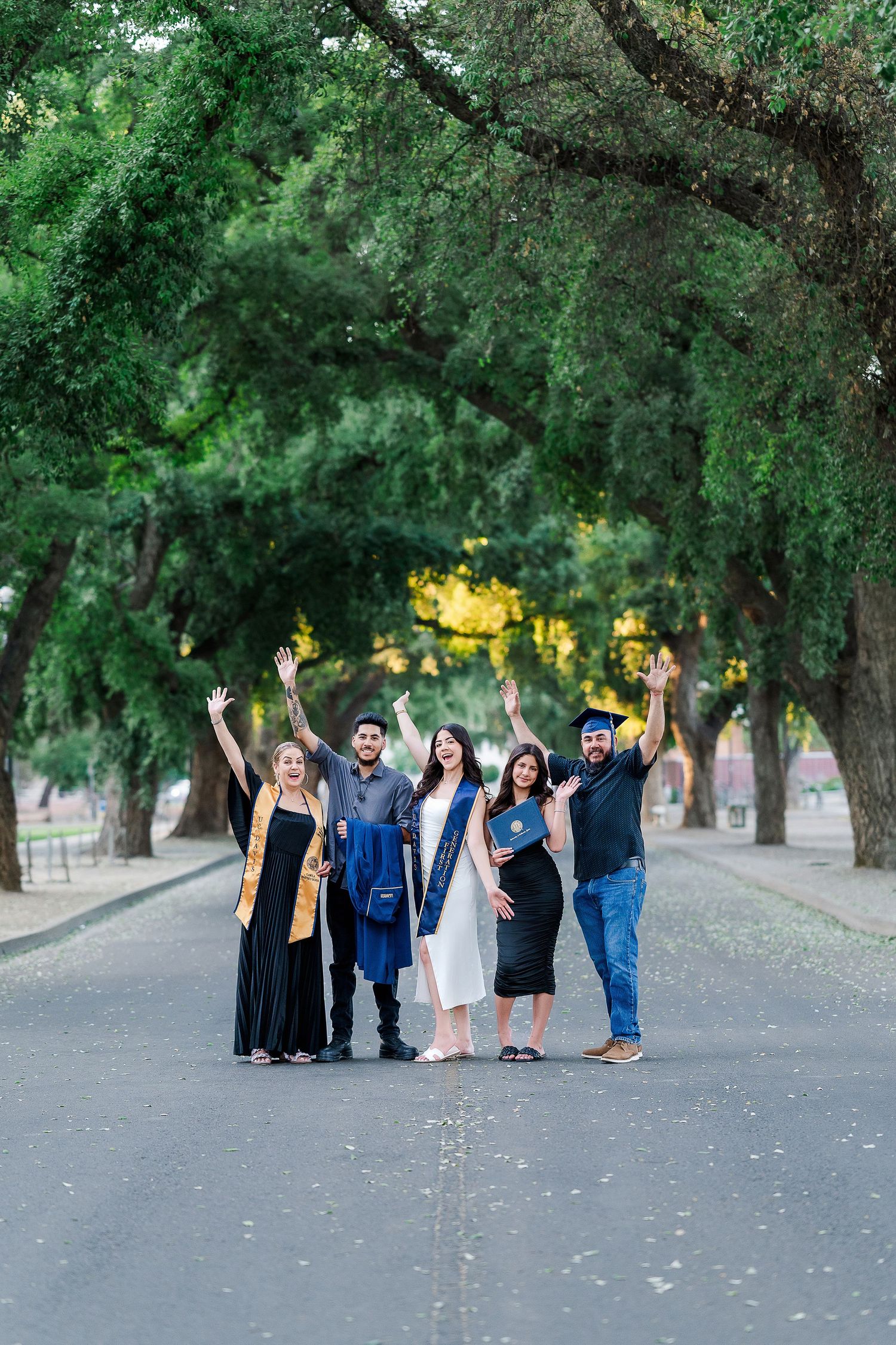Group of graduates in caps and gowns celebrating together on a tree-lined path at UC Davis.