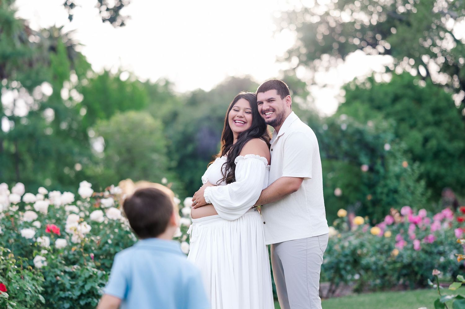 Pregnant couple embracing in a rose garden at sunset, with a young boy standing in the foreground. McKinley Rose Garden