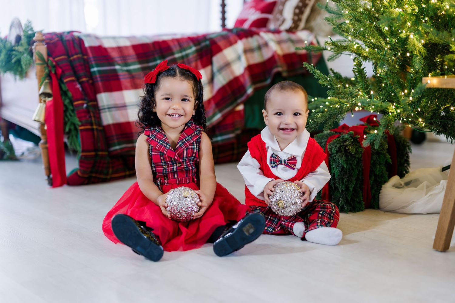 Two children sit on a white floor in matching red and plaid holiday outfits near a decorated Christmas tree.