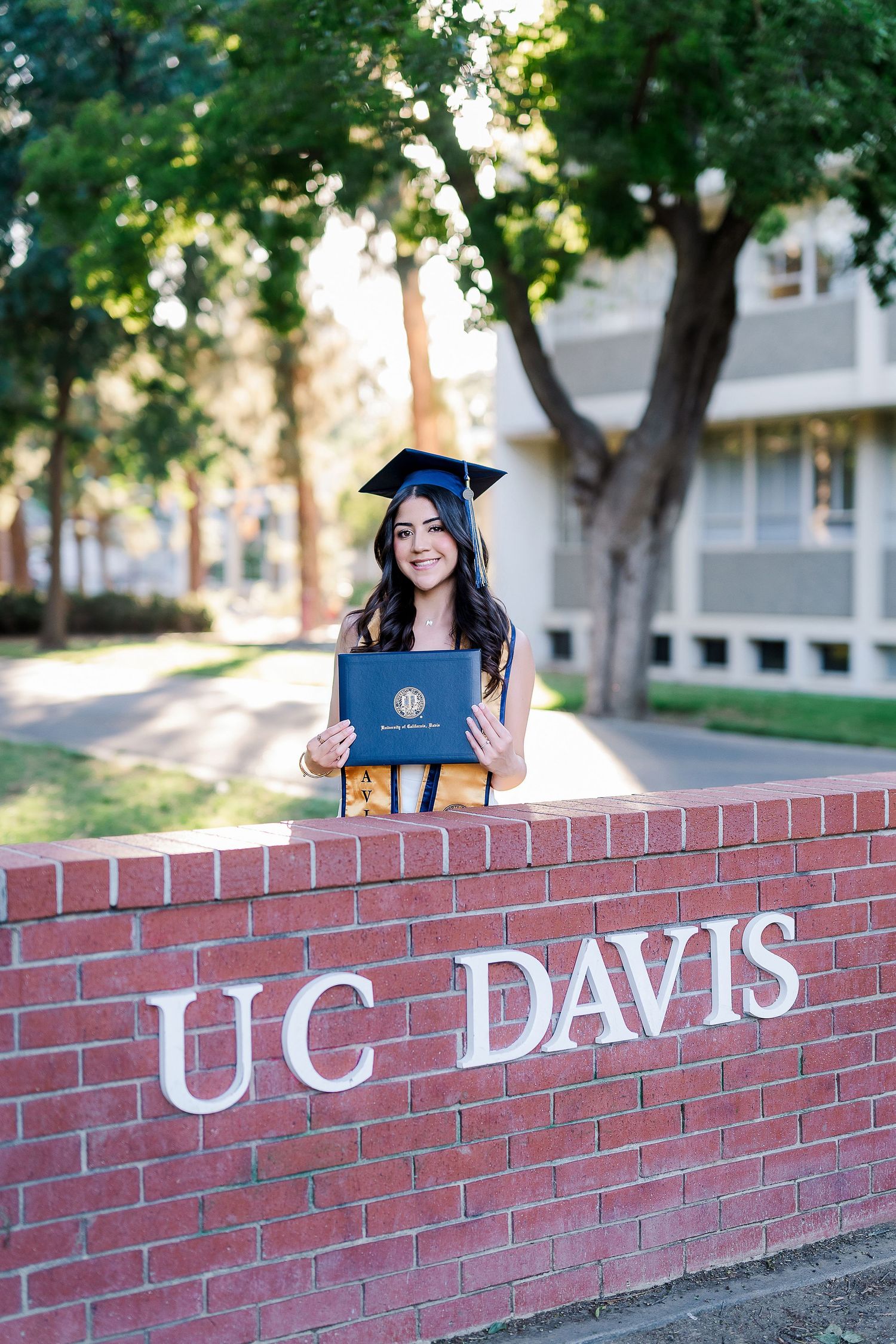 Graduate displaying diploma while posing on UC Davis campus brick wall.