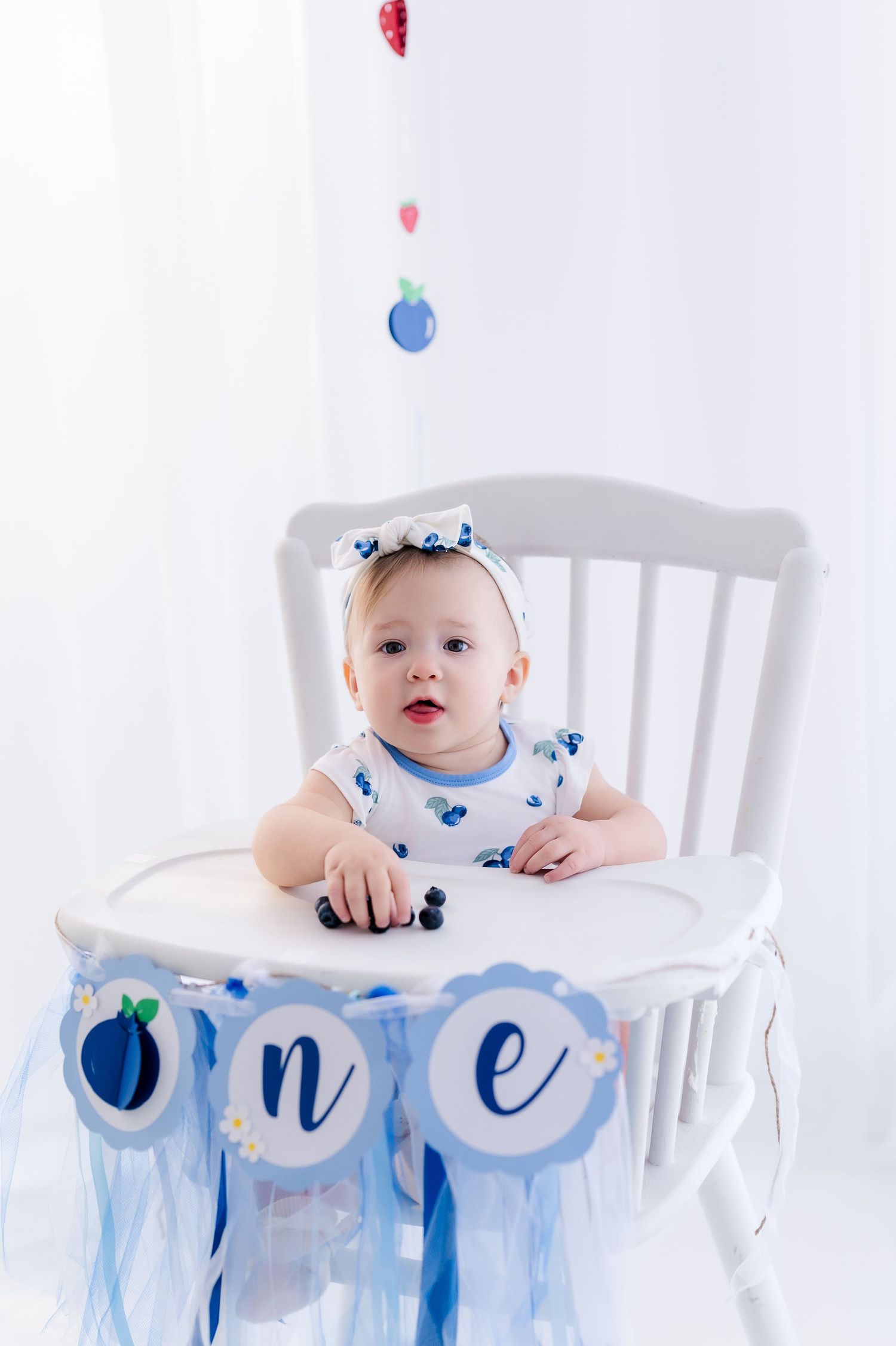 Baby girl in a white high chair celebrating her first birthday with blueberry decorations and an 