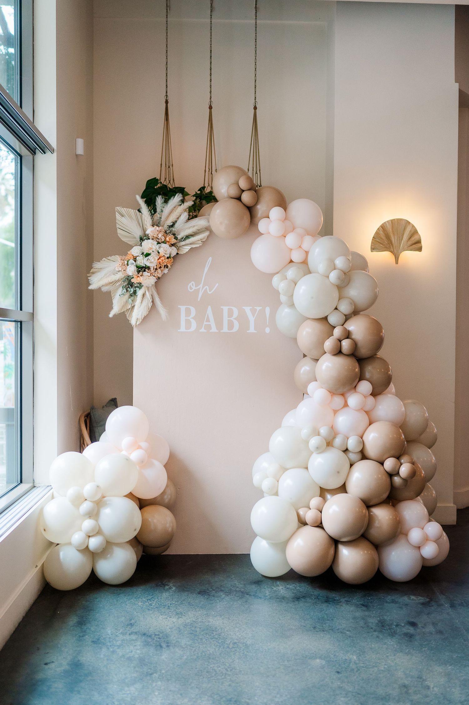 Elegant baby shower decor featuring white balloon arch and tropical leaf arrangements against beige wall.
