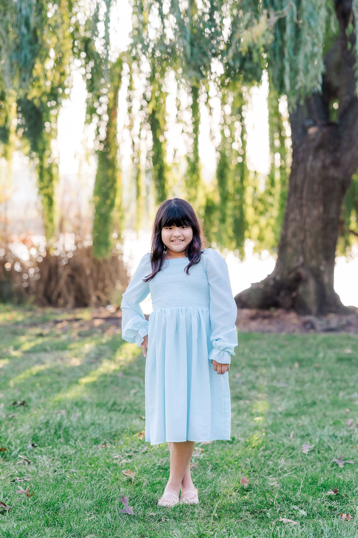 A young girl in a light blue dress plays in a sunlit park with weeping willow trees in the background.
