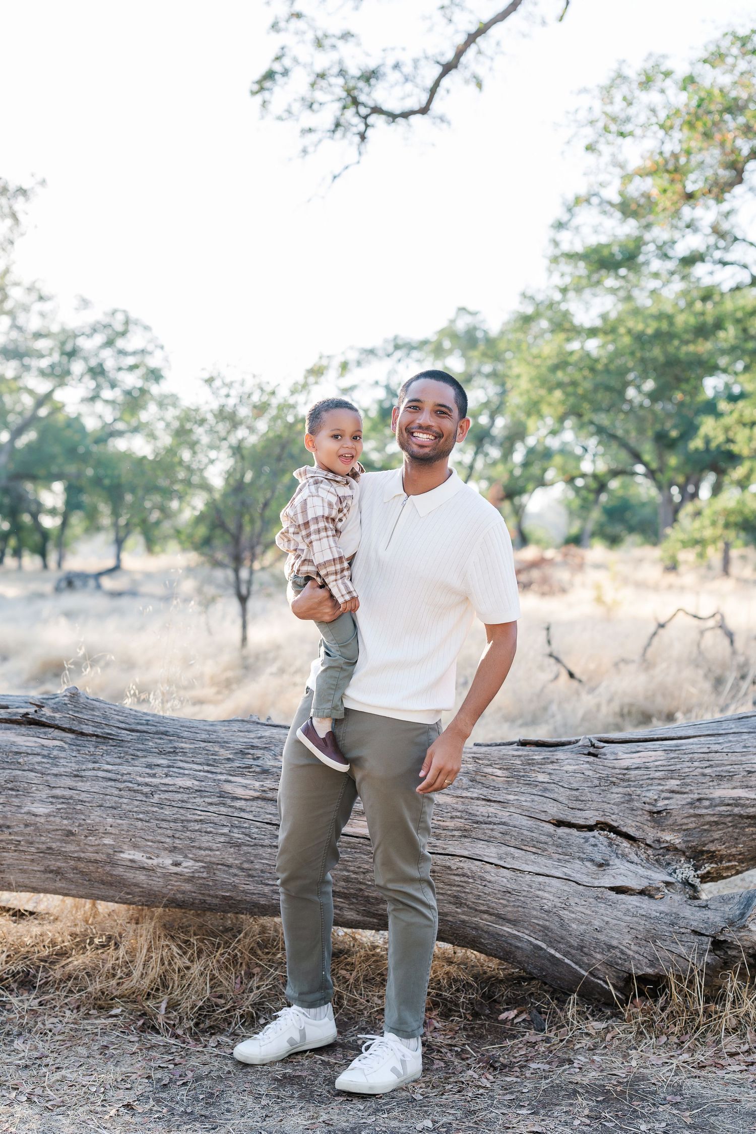 A parent and child stand together near a fallen log in a dry grassy field during golden hour.