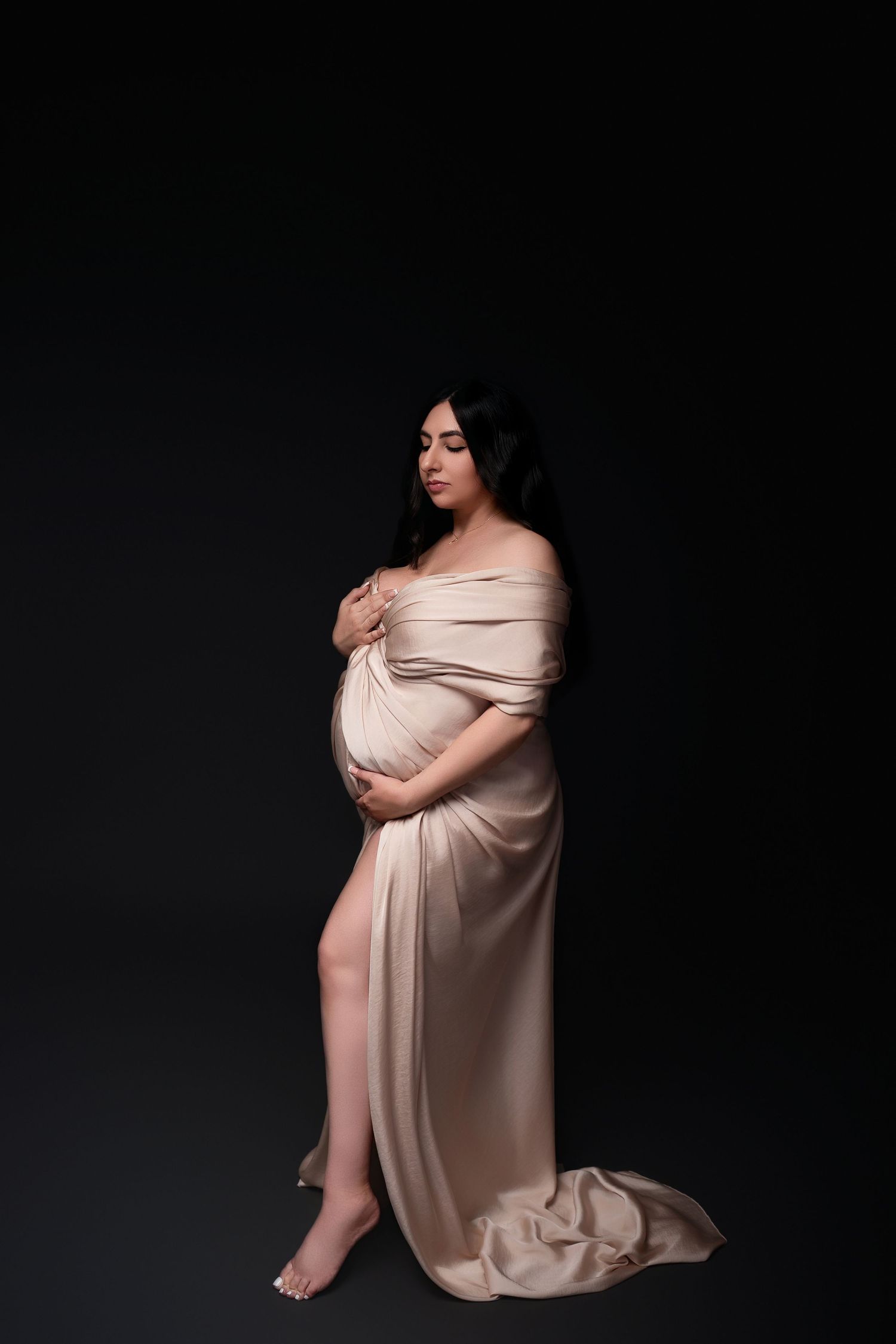 Elegant maternity portrait with woman in flowing champagne dress against dark background.