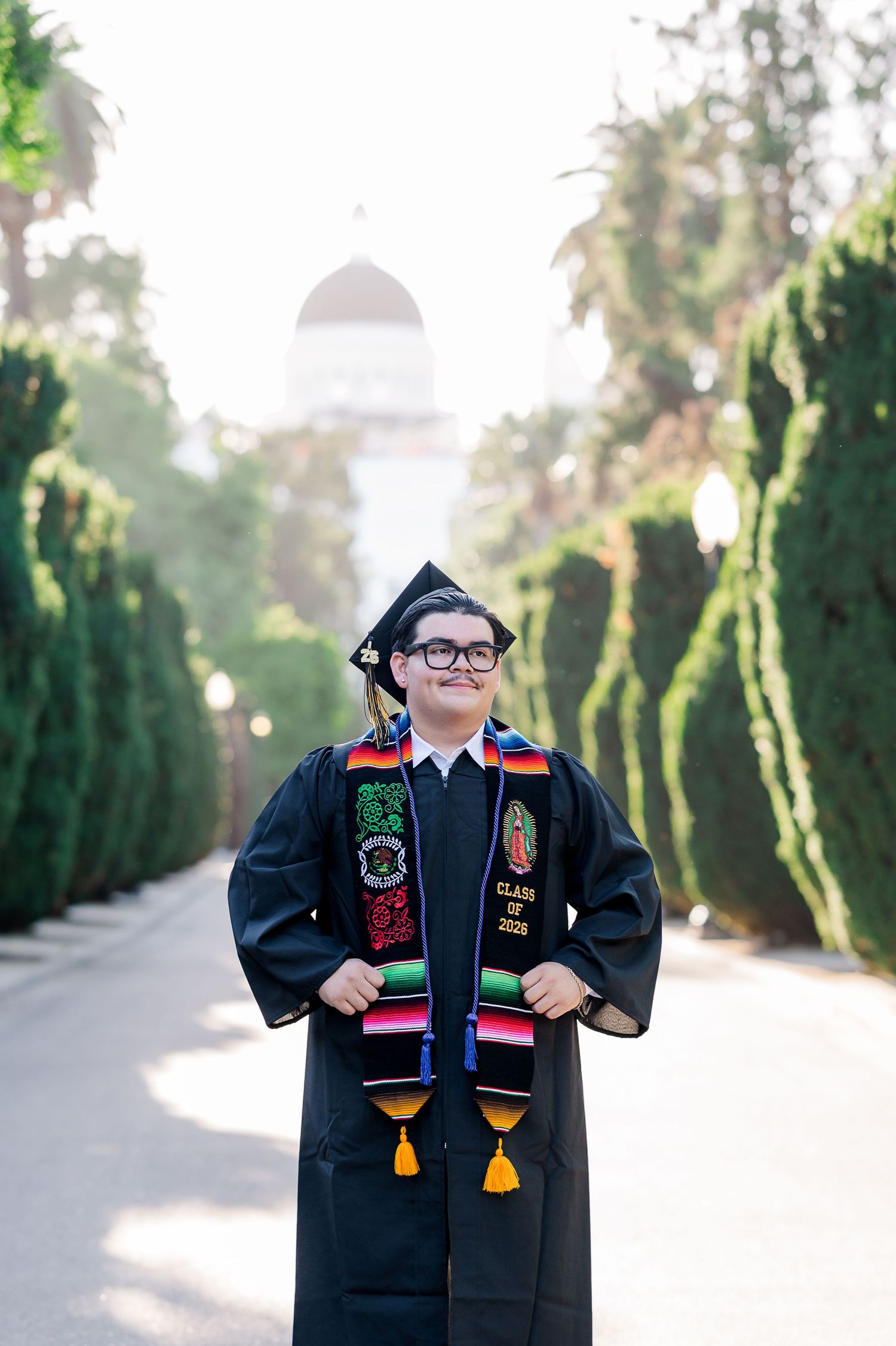 Graduate in black cap and gown with colorful sash poses proudly outdoors near a domed building and lush green hedges.
