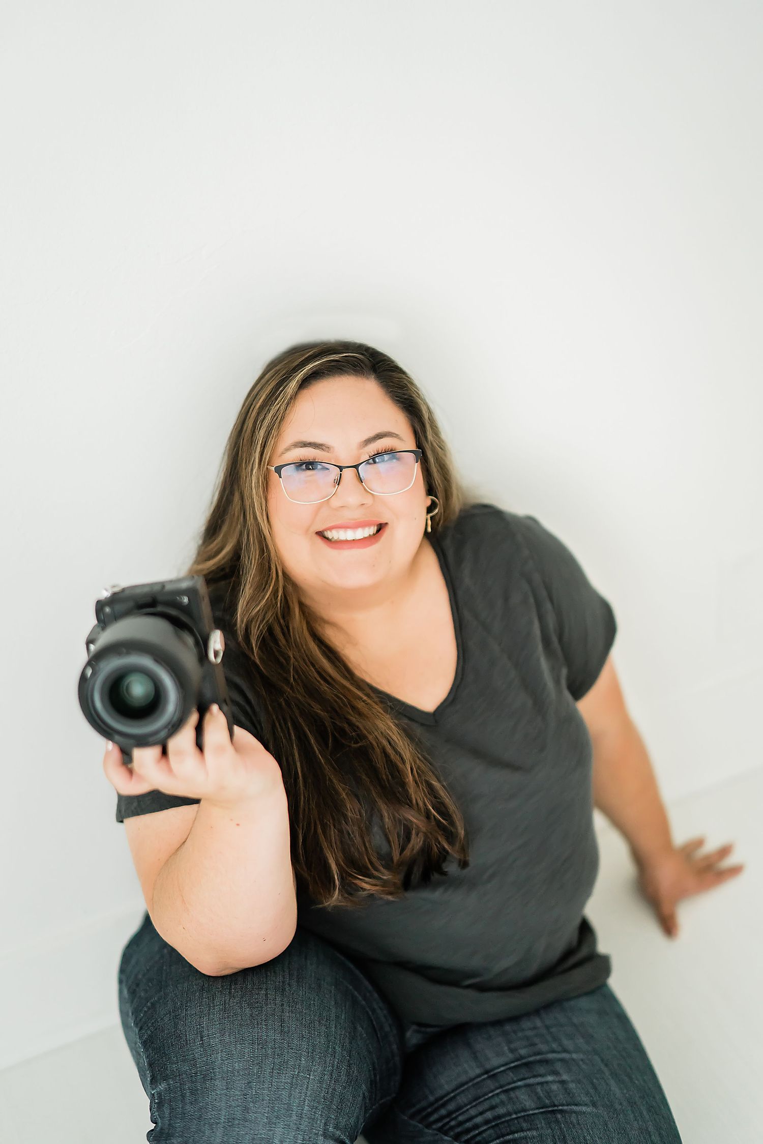 Smiling woman with glasses holding a DSLR camera, wearing a gray t-shirt and jeans, seated against a white wall. Amy Medina Photography