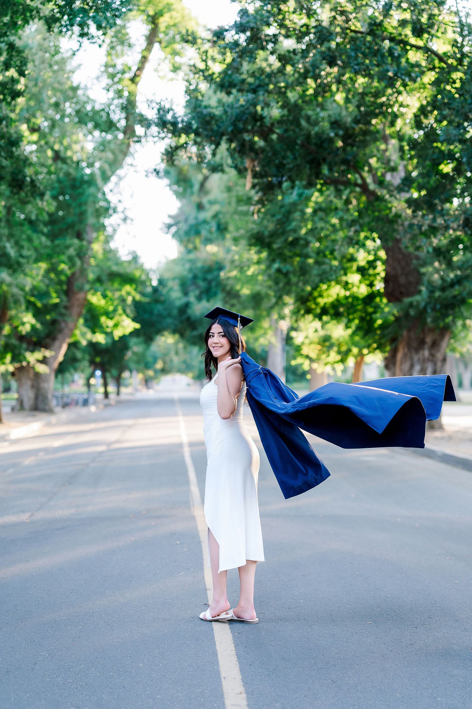 Graduate in white dress and blue cap strikes playful poses on empty road surrounded by trees.