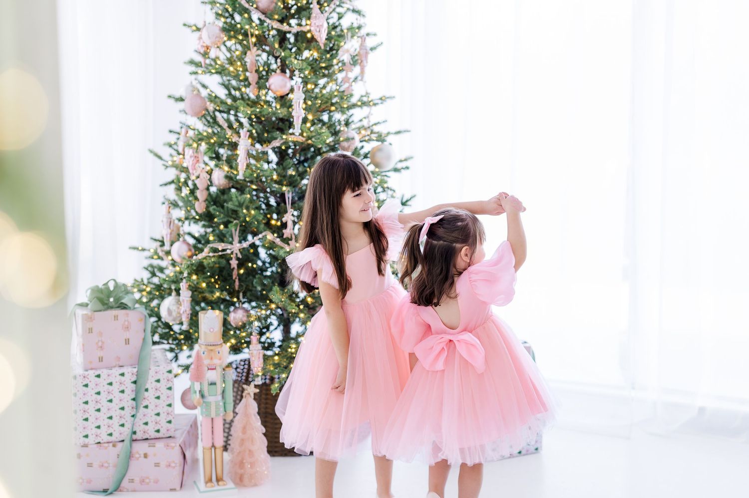 Two figures in pink tulle dresses dance together in front of a decorated Christmas tree in a bright white room.