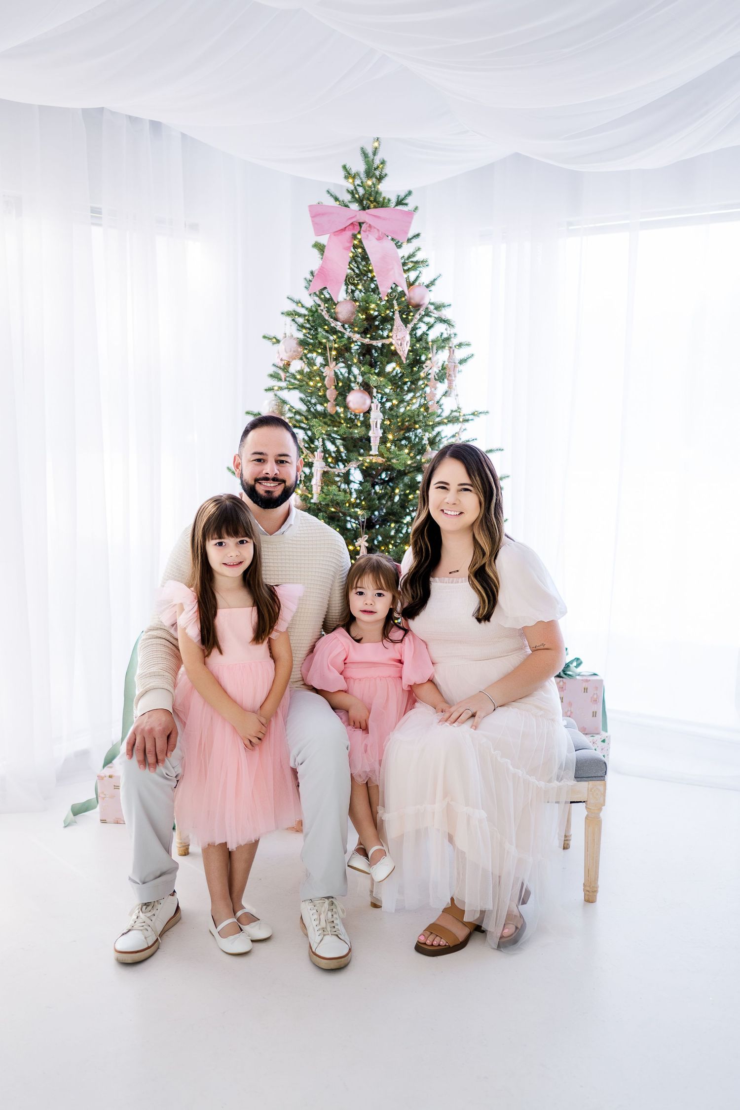 A family poses together in front of a decorated Christmas tree with white draping and pink accents.