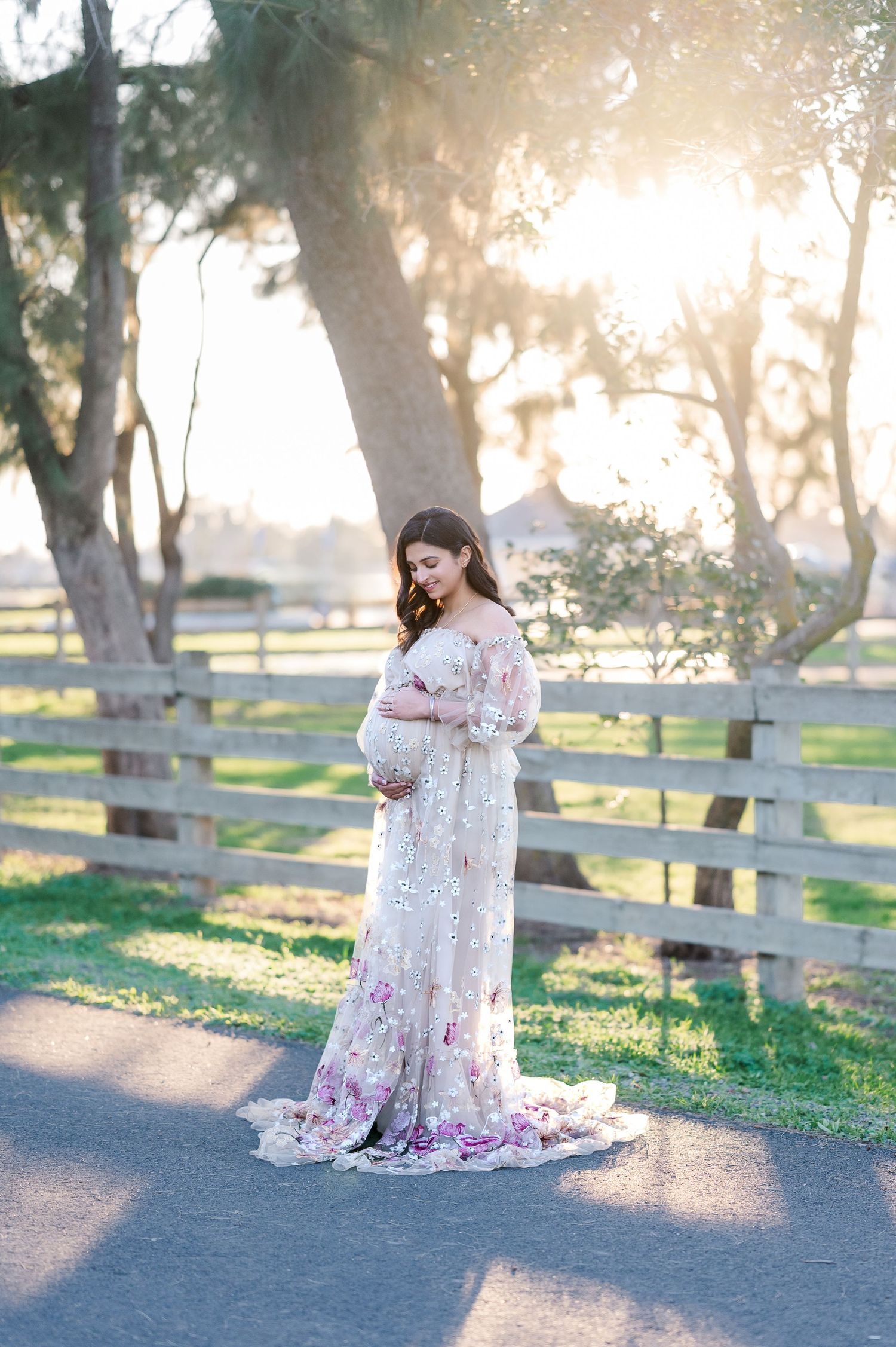 Floral maternity dress photoshoot in a sunlit park with wooden fences and trees at golden hour.
