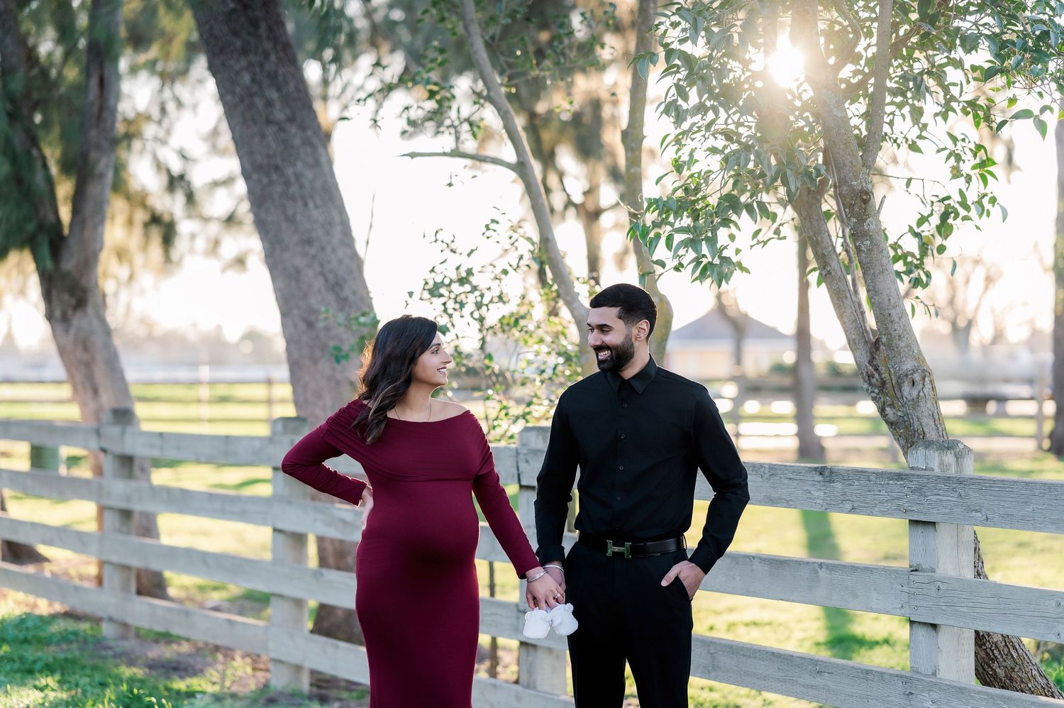 A couple shares a romantic moment together in a sunlit park with wooden fences and trees in the background.