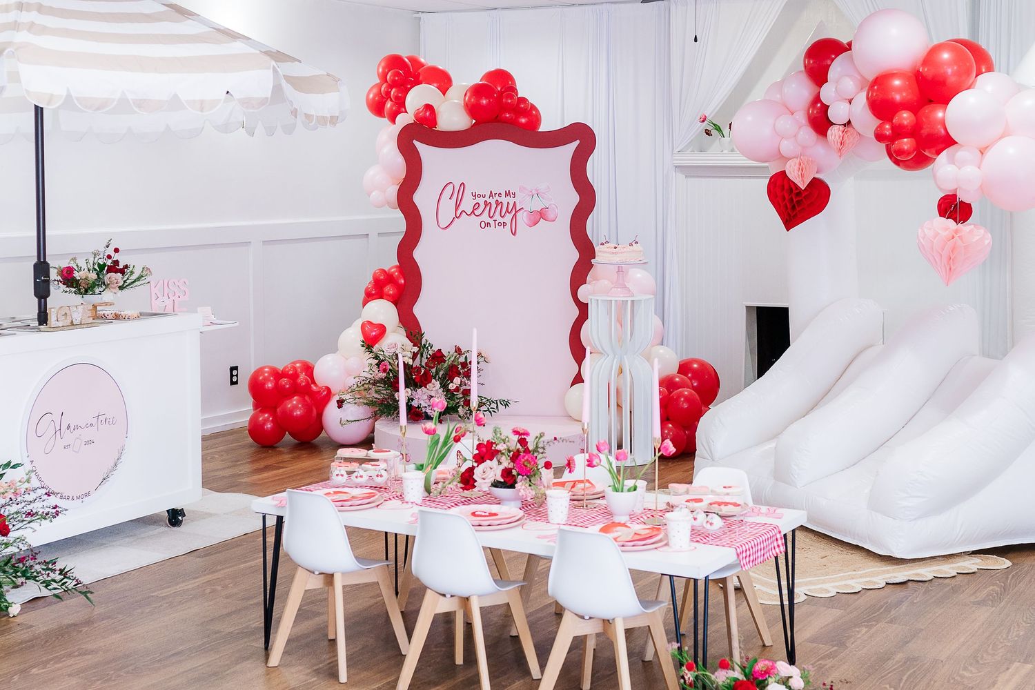 Valentine's day party setup with red and pink balloons, white chairs, and festive table decorations in a bright room.
