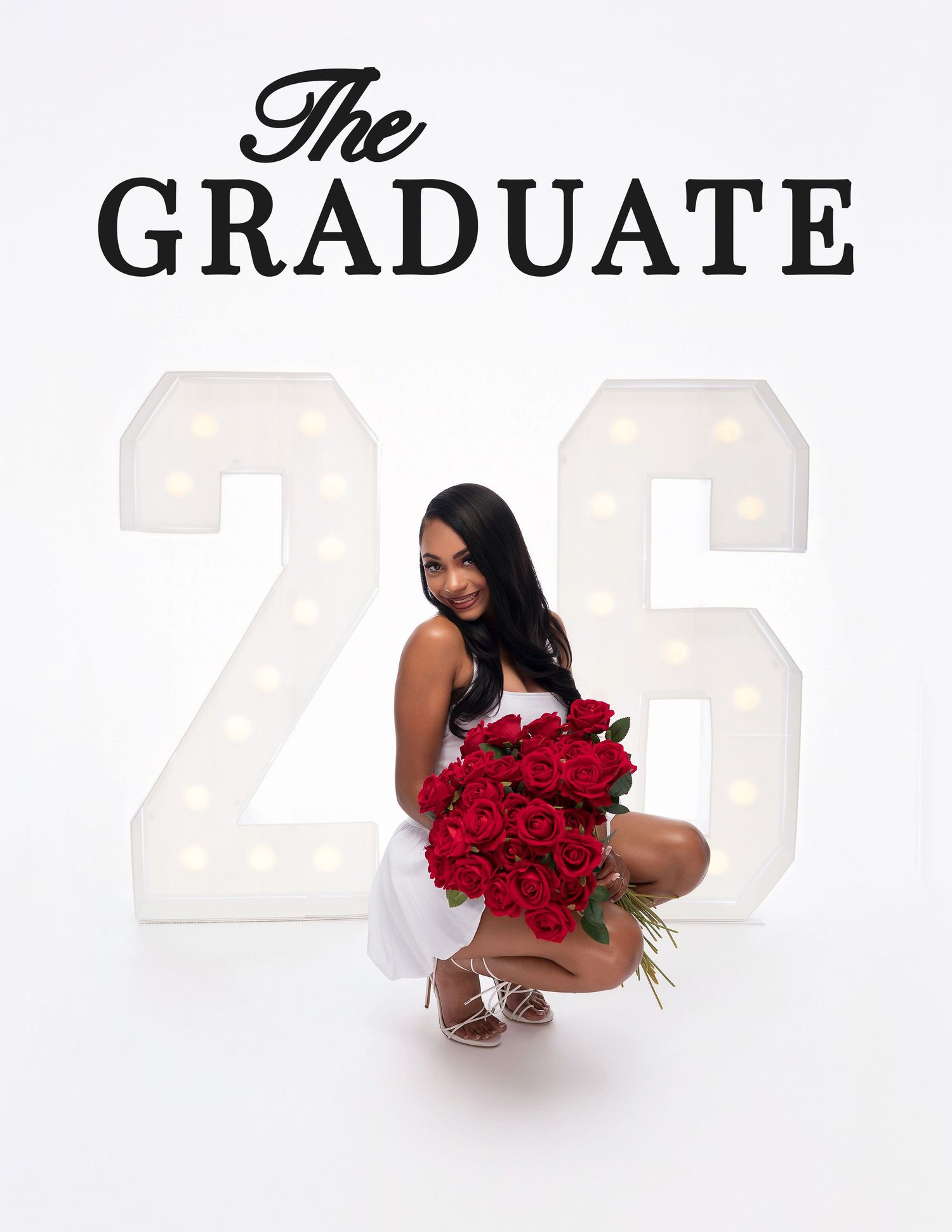 A graduate poses with red roses bouquet in front of illuminated 26 marquee letters on white background.