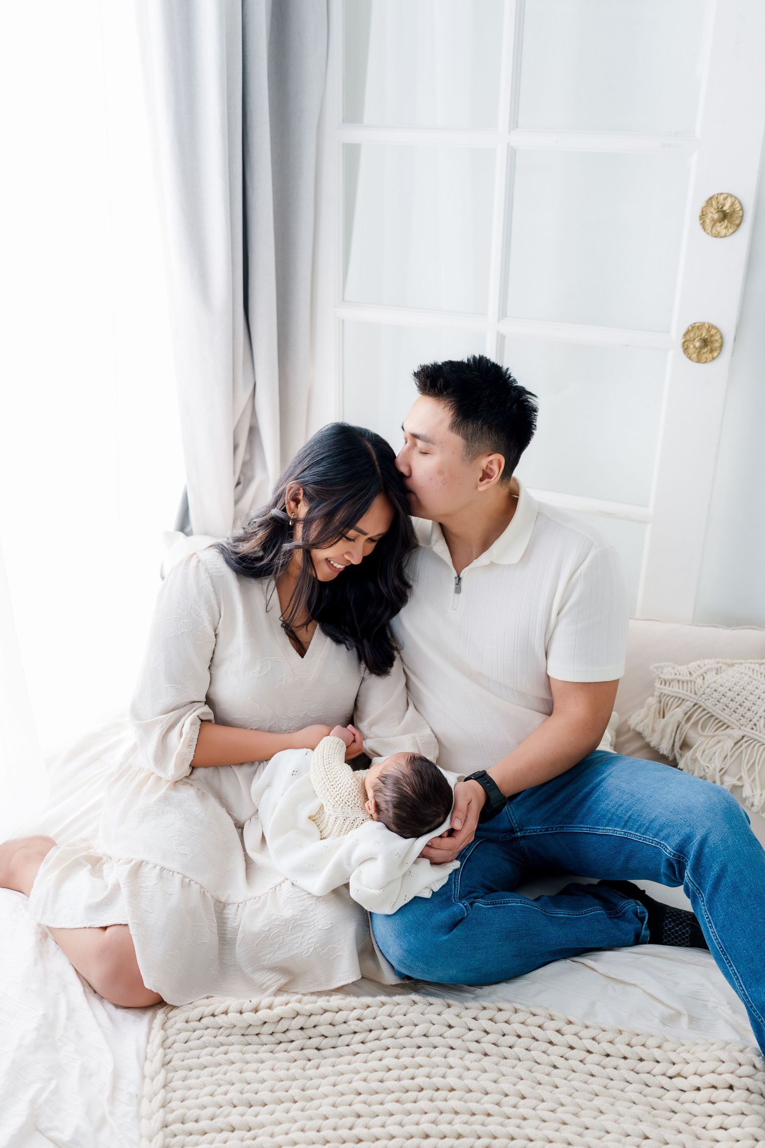 A couple sits on a bed, lovingly gazing at their newborn baby, with the father kissing the mother's head. Sacramento Newborn Photographer