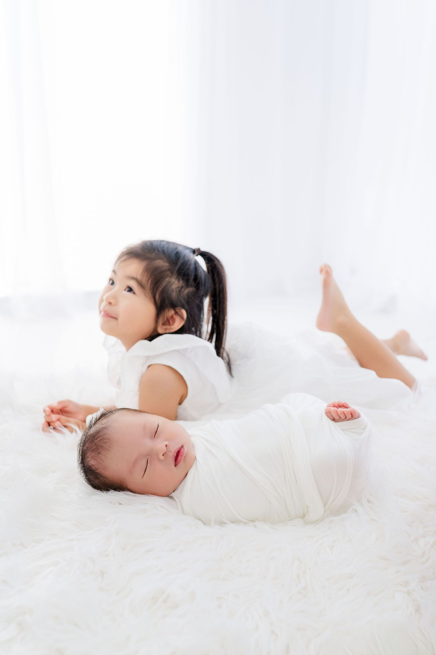 A young girl in white lies on a fluffy white blanket beside a sleeping swaddled newborn baby.