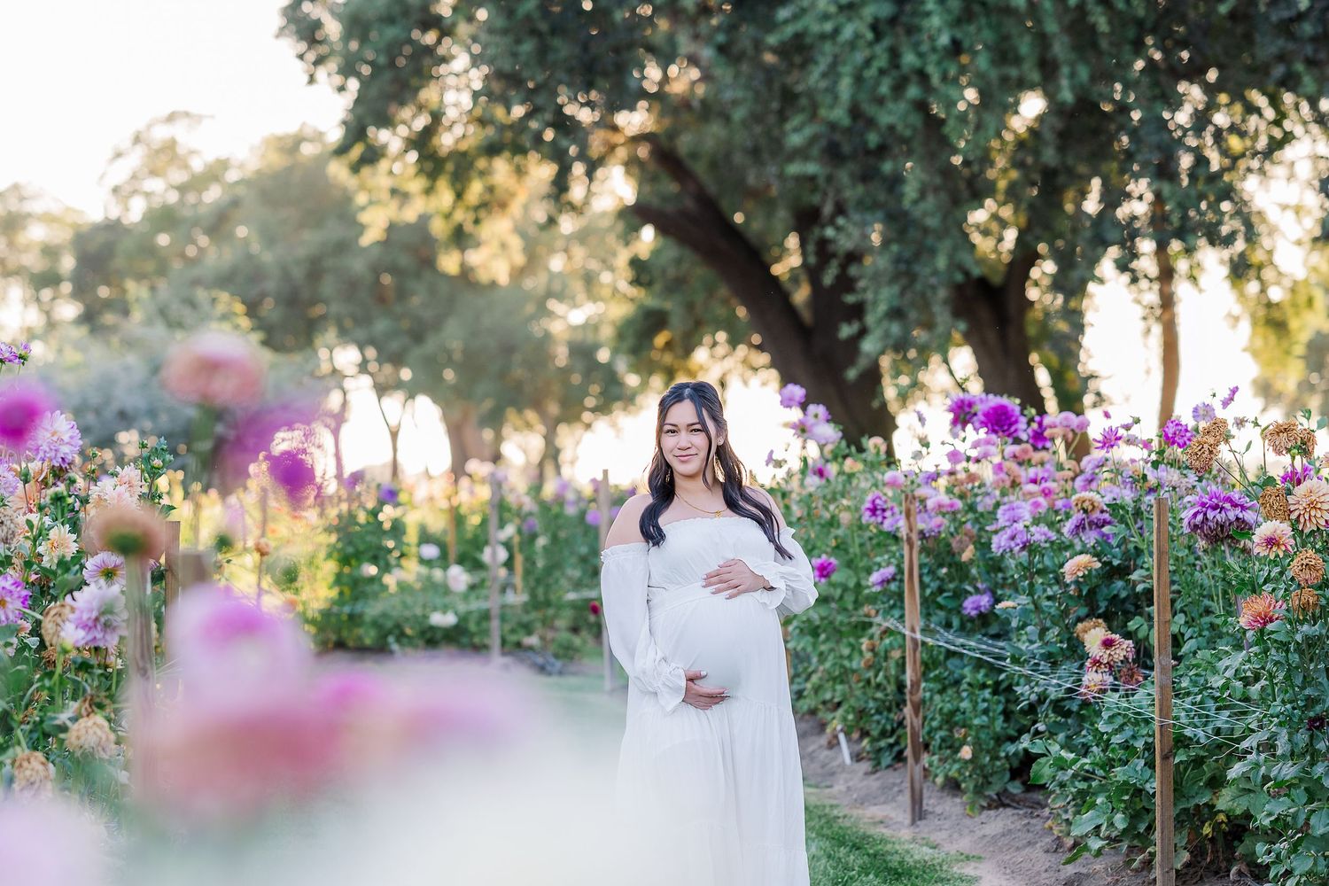 Pregnant woman in white dress smiling among colorful flowers in a sunlit garden during a maternity photo session.