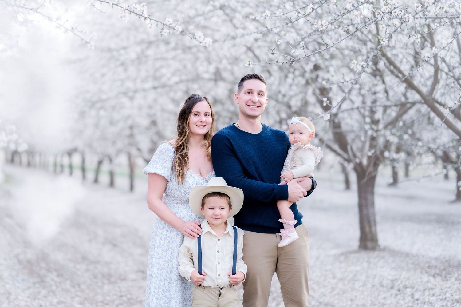 Family of four poses among blooming white trees, parents smiling with toddler boy in suspenders and baby in arms.