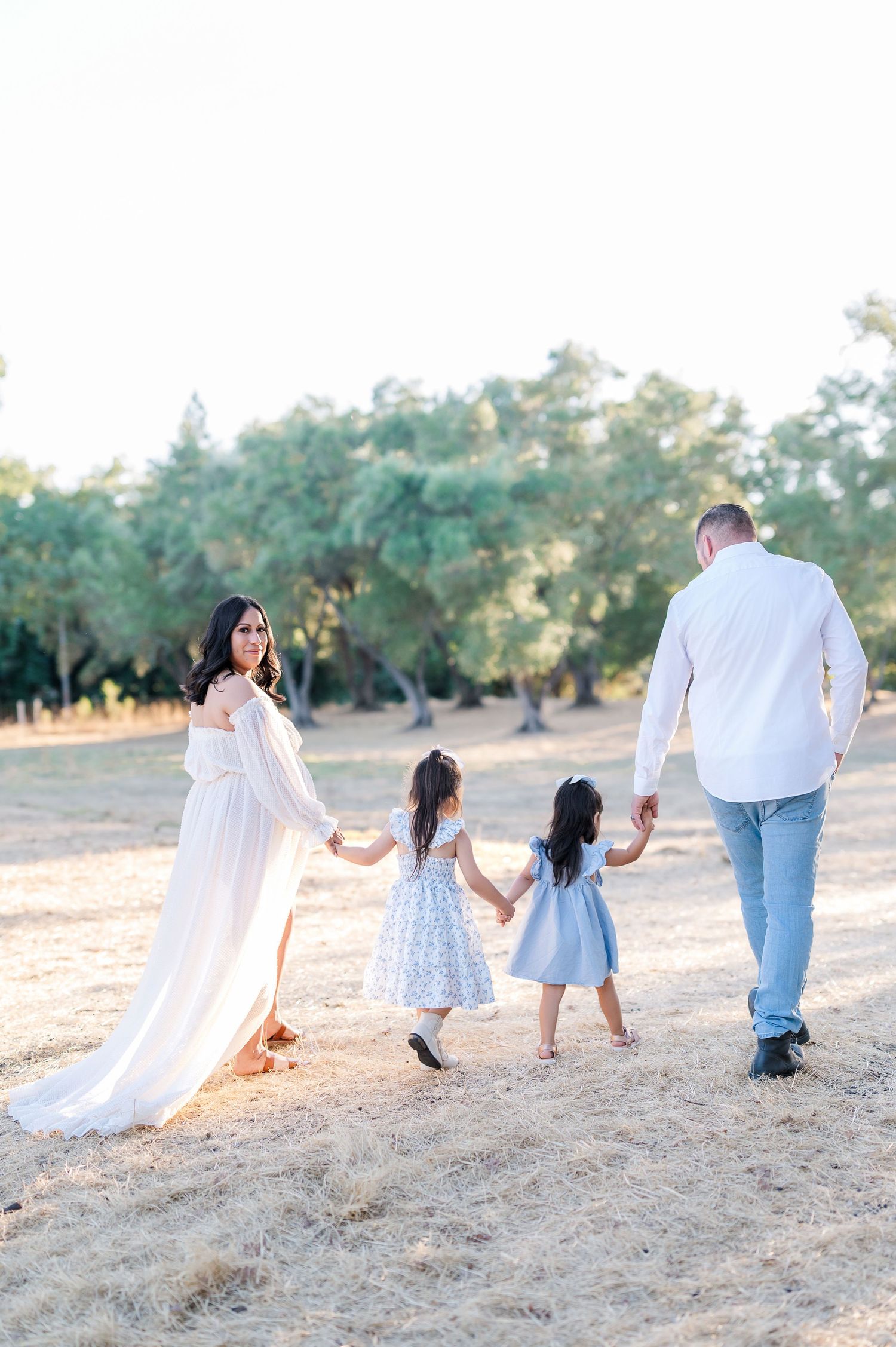 Family of four walking hand-in-hand through a sunlit field, mother in white gown, father in white shirt and jeans.