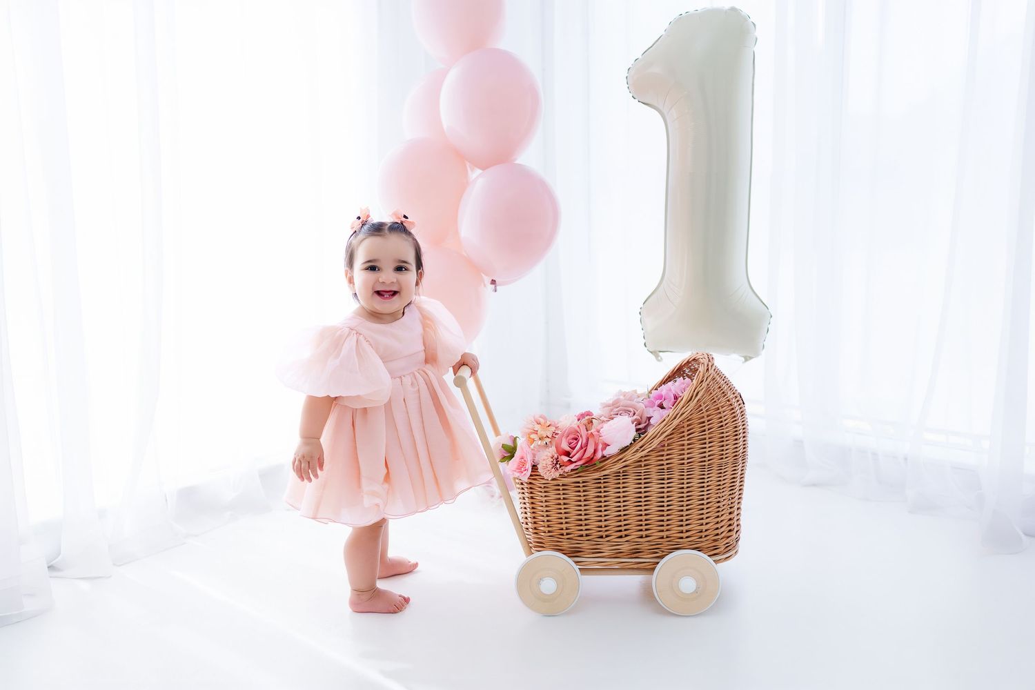 Baby girl in pink dress smiles beside a wicker pram, pink balloons, and a number one balloon for her first birthday.