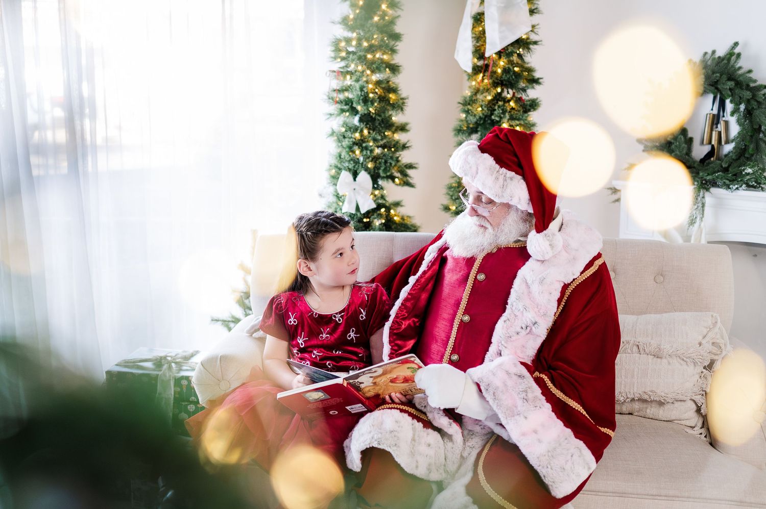 A festive Christmas scene with Santa Claus sitting in a chair near a decorated tree while interacting with a young visitor.