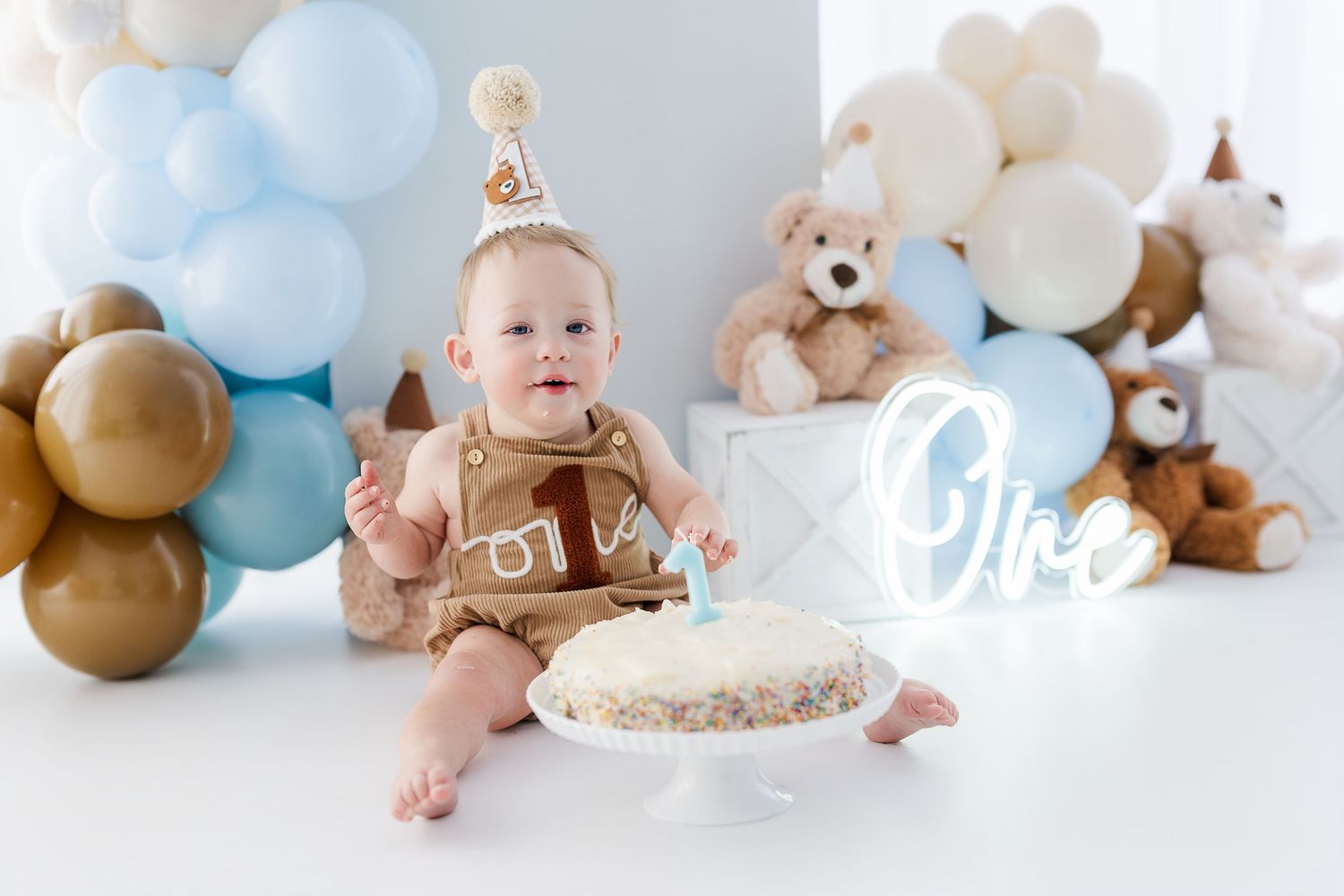 Baby's first birthday cake smash with blue and gold balloons, teddy bears, and an 'One' sign in a white studio.