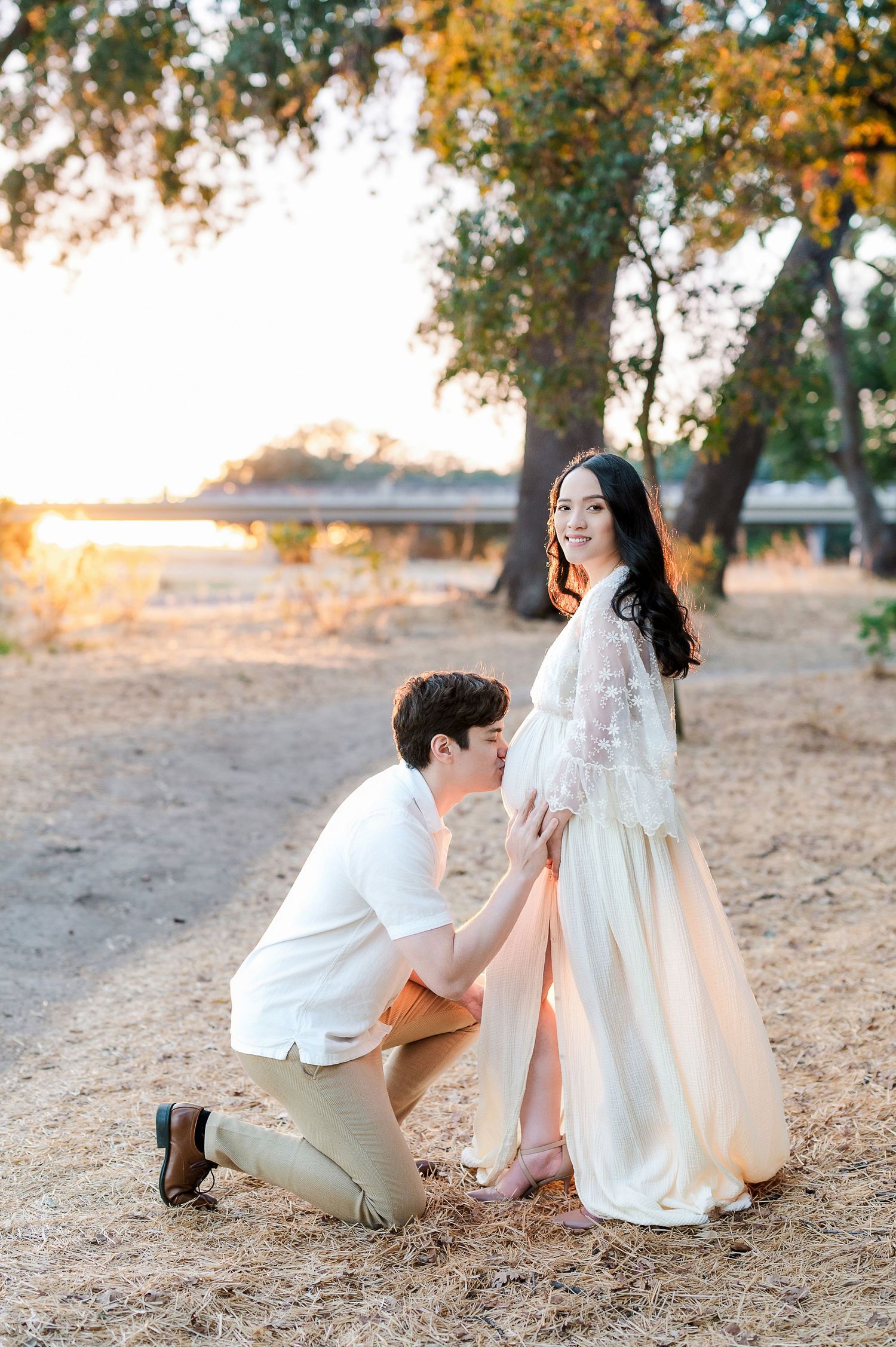 A man kneels kissing a pregnant woman's belly outdoors at golden hour, surrounded by trees near a lake.