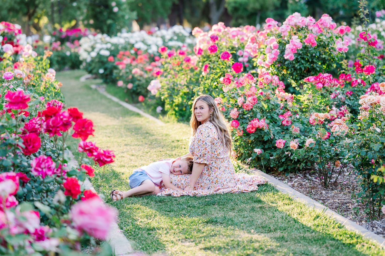 A woman in a floral dress sits gracefully in a blooming rose garden.