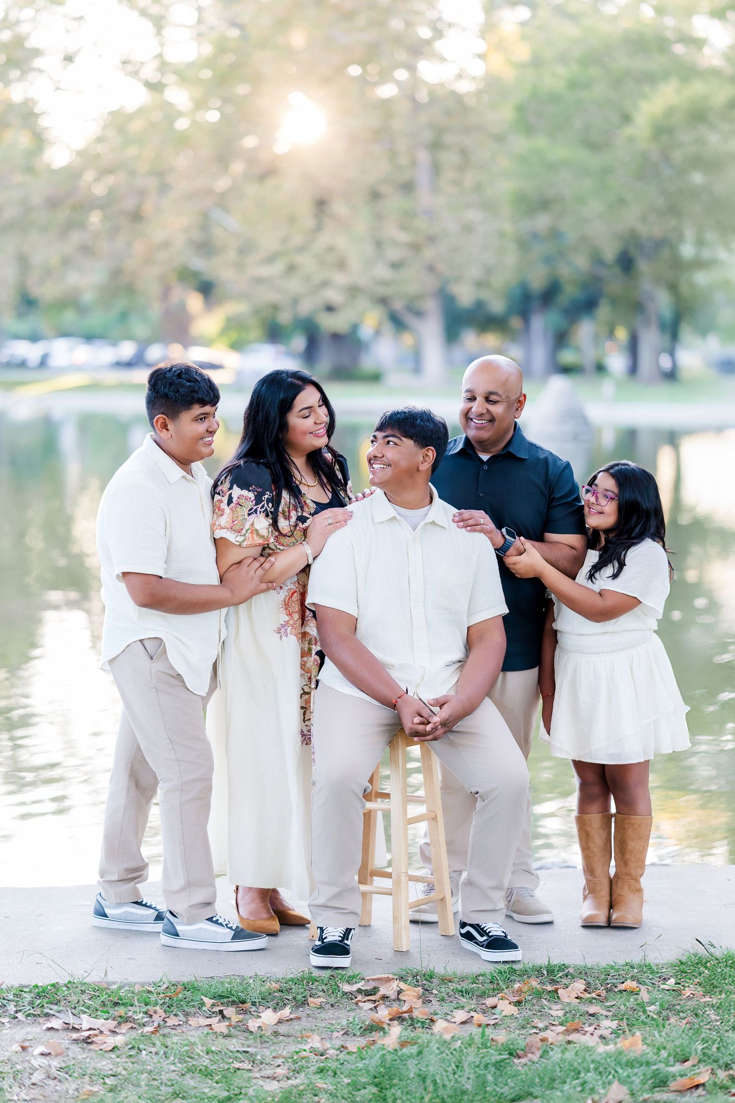 Family photo session by a lake with people wearing coordinated white and beige outfits sitting on wooden chairs.
