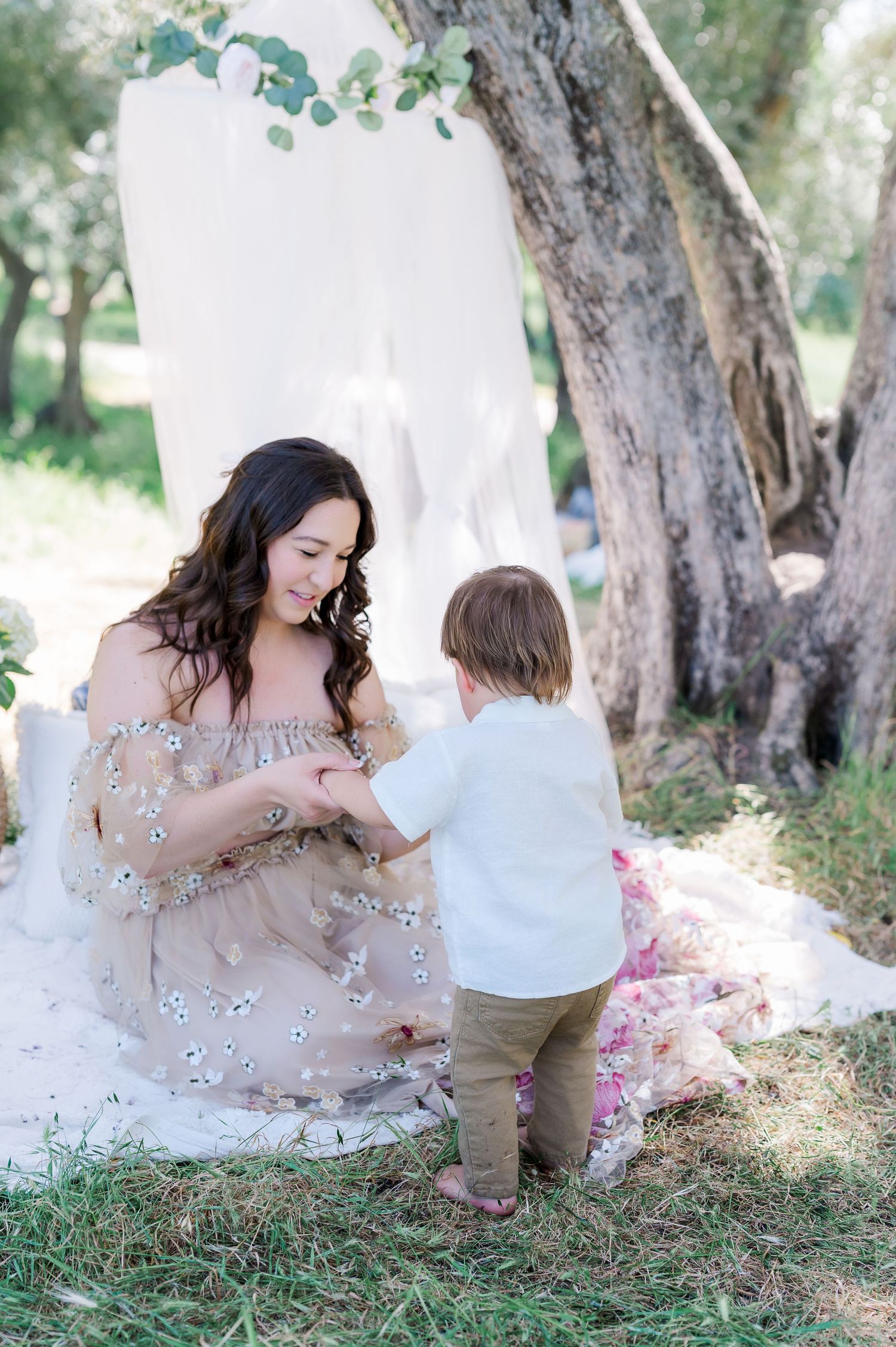 A mother and child share a tender moment together under olive trees in a dreamy outdoor setting.