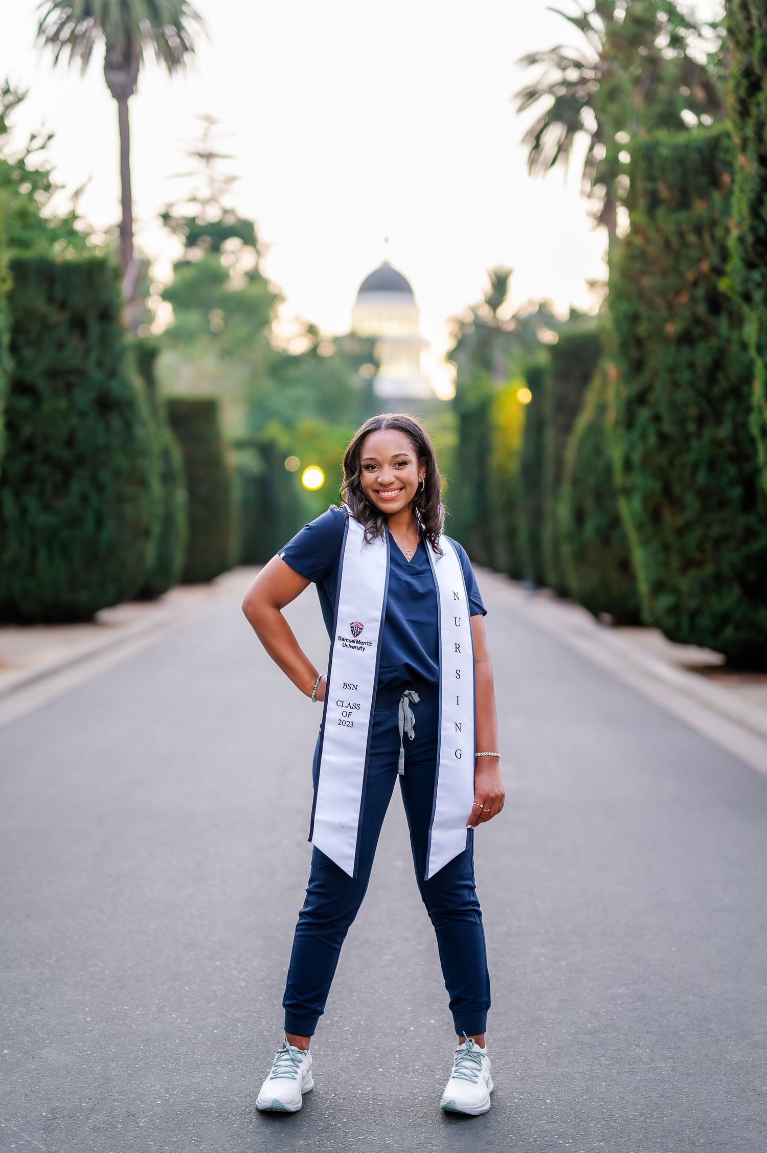 Graduate posing confidently on palm tree-lined path in graduation attire.