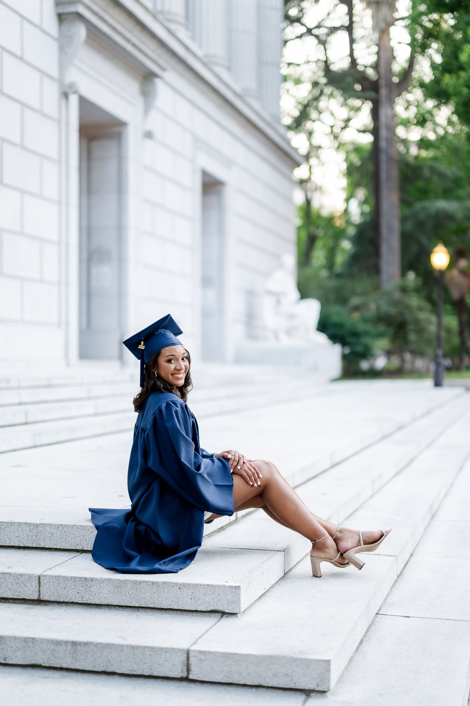 Graduate in blue cap and gown poses on white stone steps.