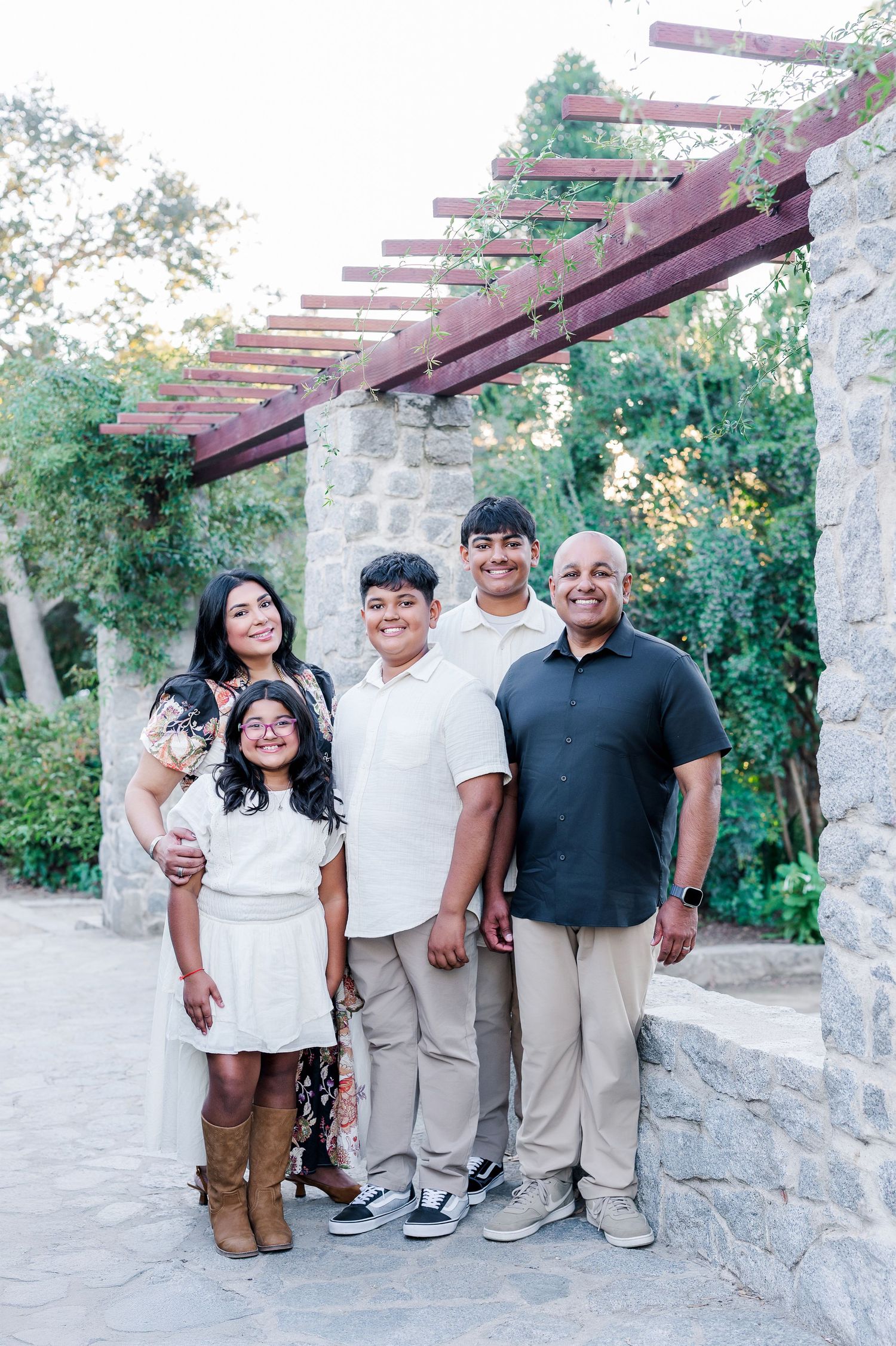 A family group poses together outdoors under a wooden pergola structure against stone walls and greenery.