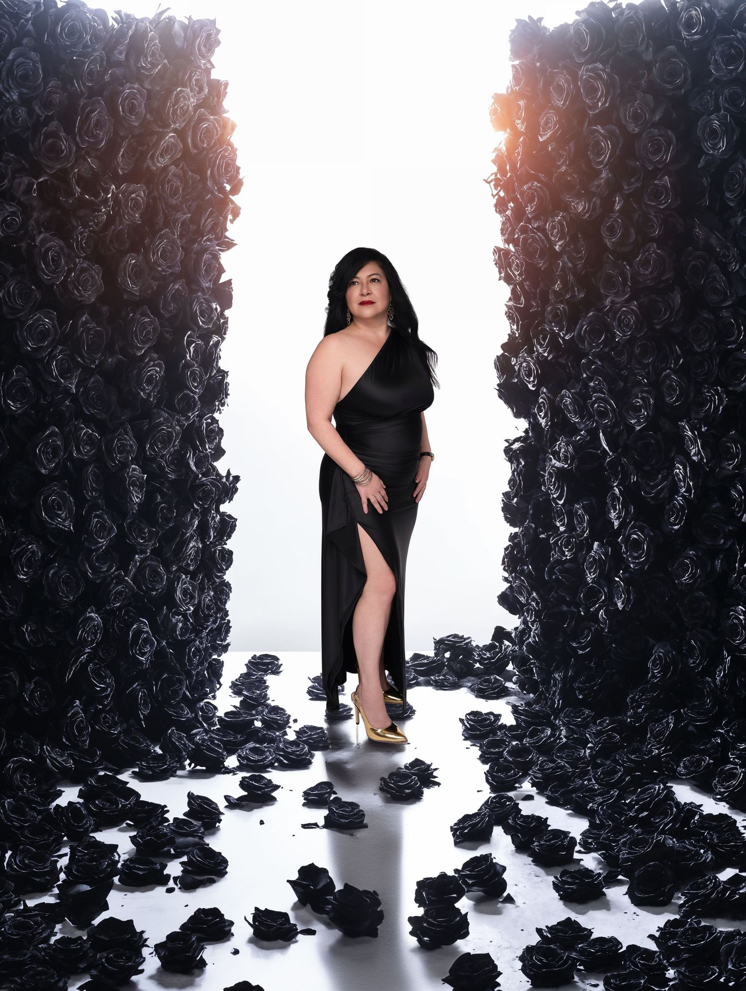 A figure in black poses dramatically against a white backdrop surrounded by scattered dark rose-like shapes on the floor.