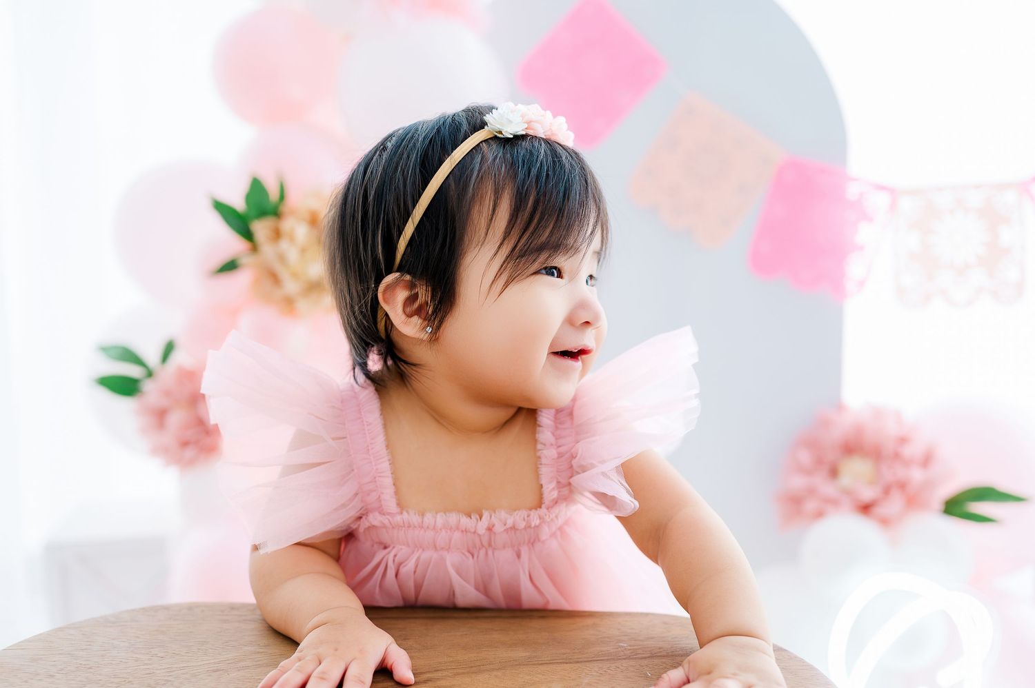 A baby girl in a pink tulle dress with a floral headband smiles at a first birthday cake smash photo session.