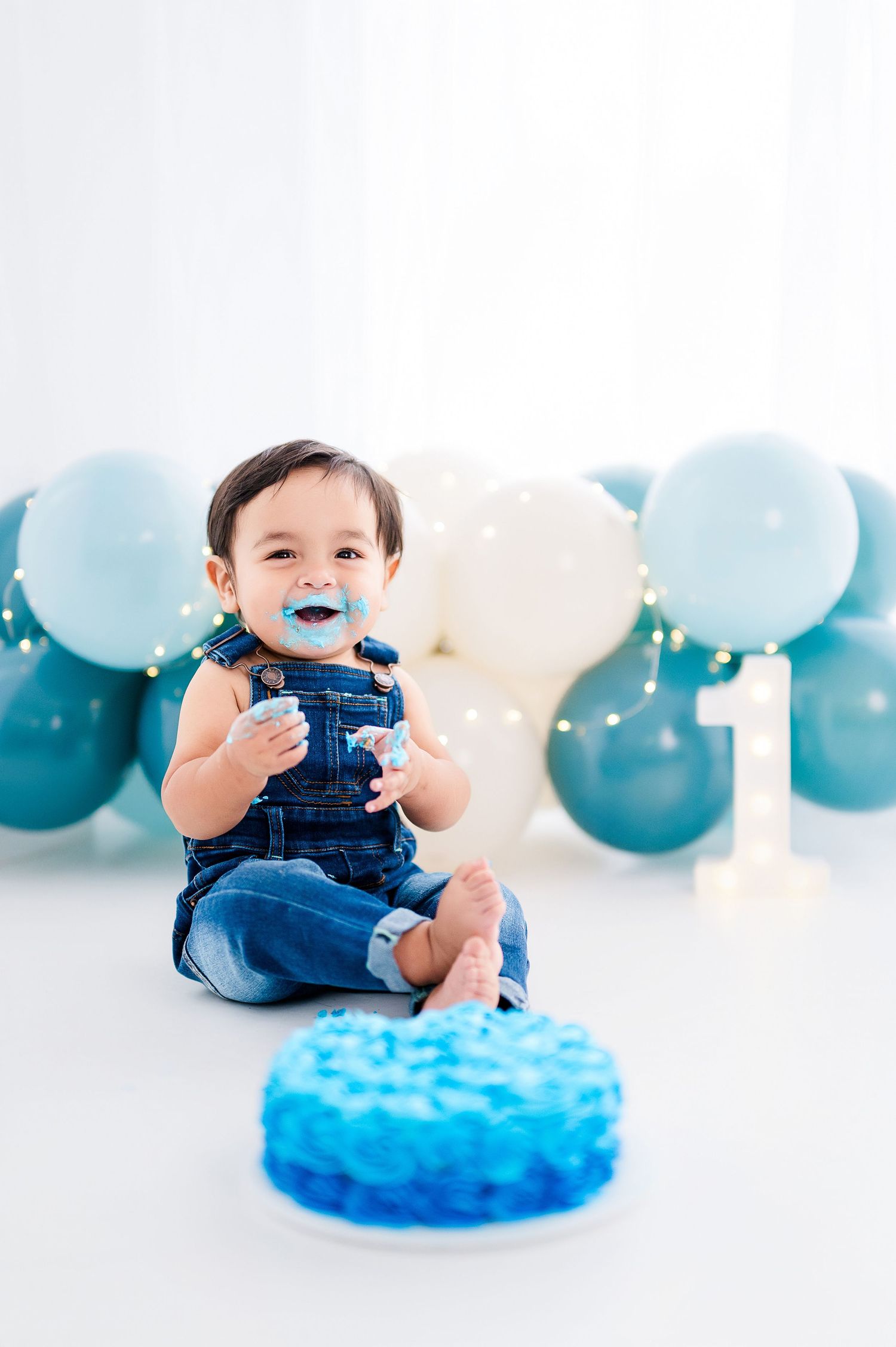 Baby in denim overalls smashing blue cake during first birthday photoshoot with teal and white balloon decorations.