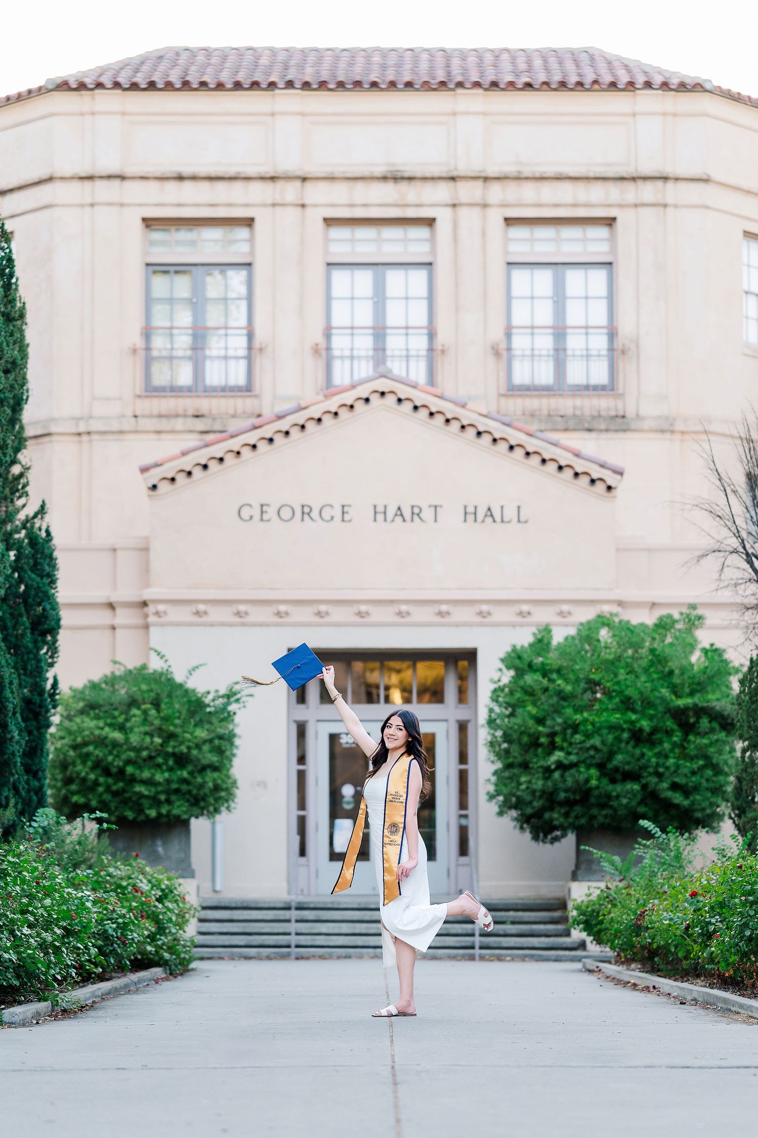 A graduate poses in front of George Hart Hall wearing academic regalia and a yellow stole.