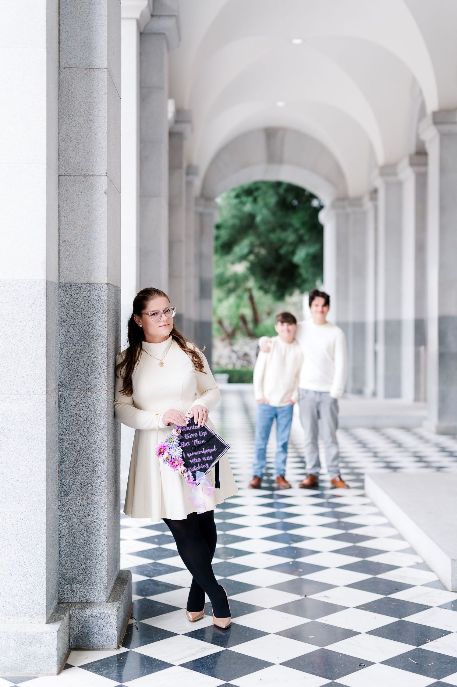 Graduate holding decorated cap in elegant arched hallway with checkered floor, family standing in background.