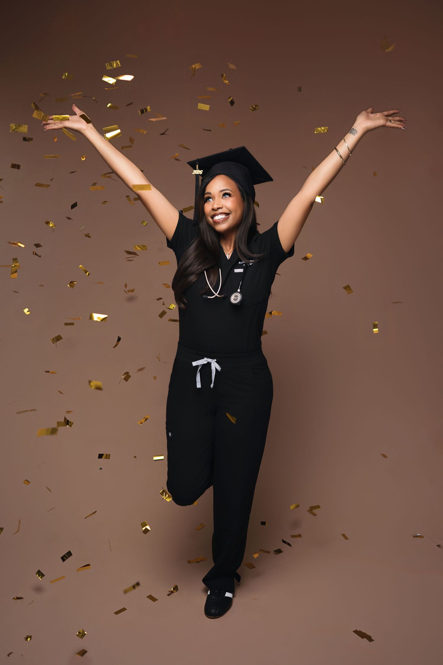 Joyful nursing graduate in black scrubs and cap celebrating with gold confetti falling around her.