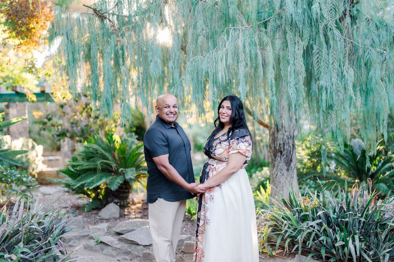 A couple poses for romantic engagement photos under a weeping willow tree in a garden setting at sunset.