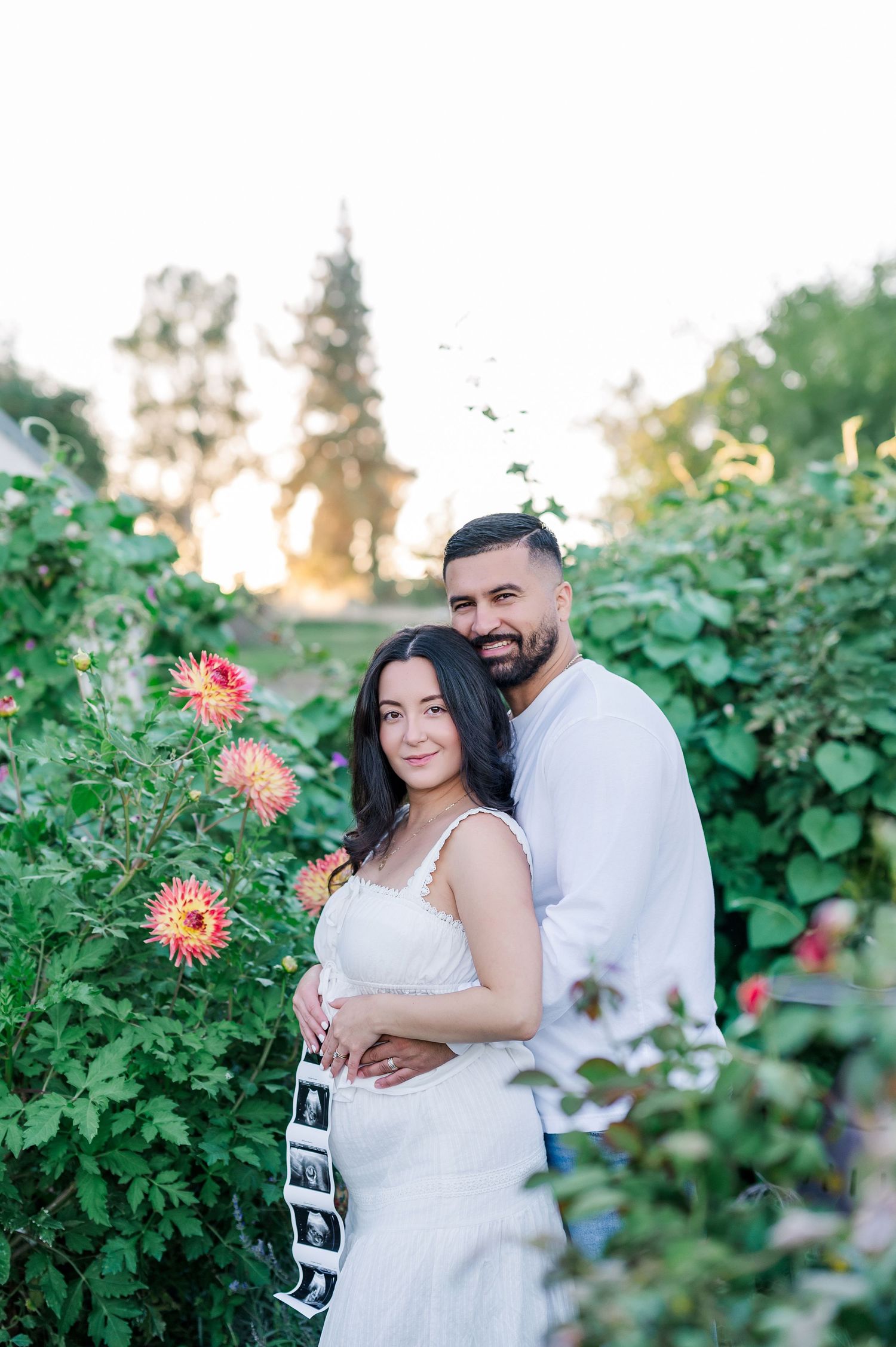 A couple shares a romantic moment in a garden filled with blooming dahlias during sunset.