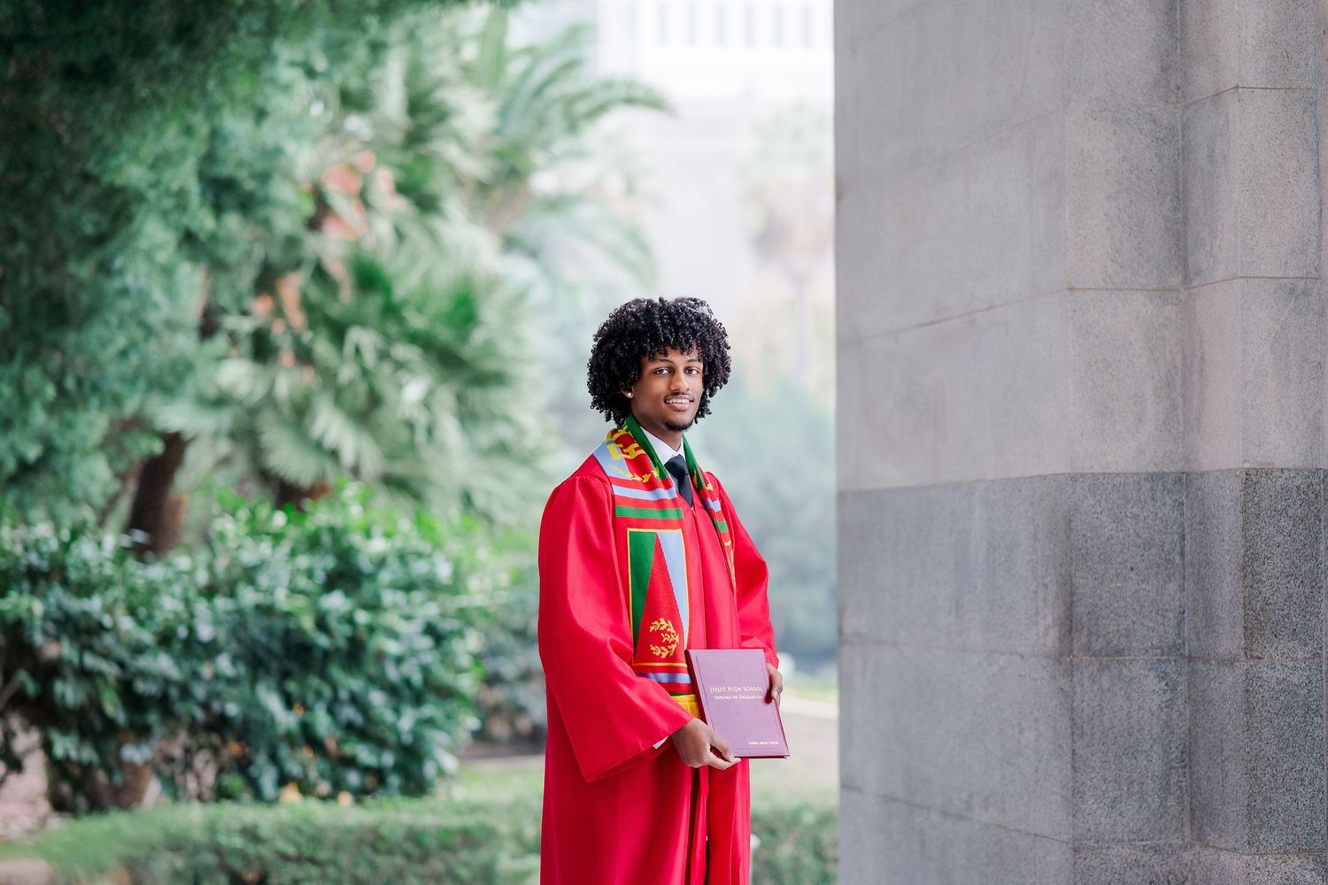 A person in a bright red coat stands in front of a stone pillar while holding a beverage on a cloudy day.