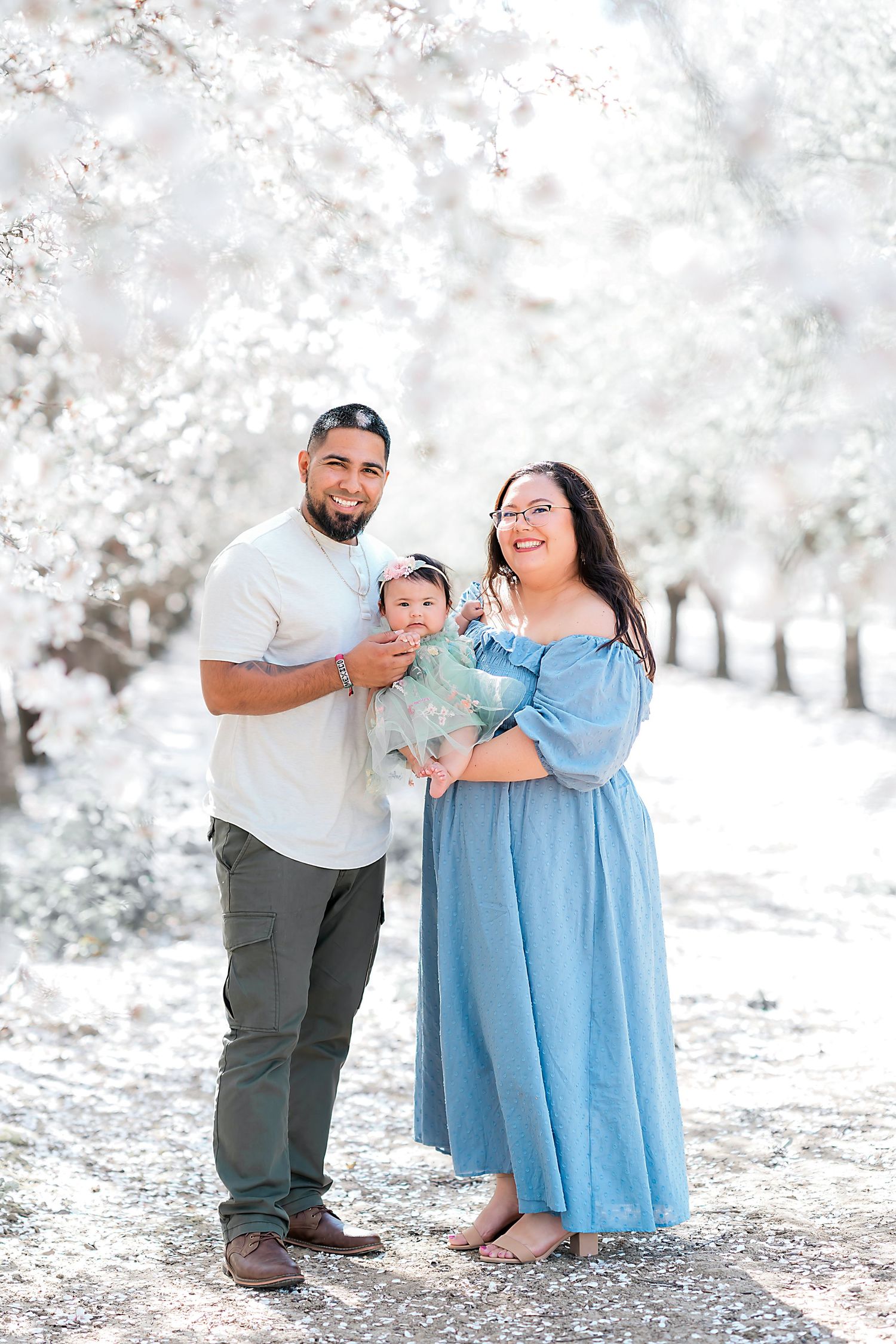 Family of three posing under blooming cherry blossoms wearing coordinated blue and white outfits.