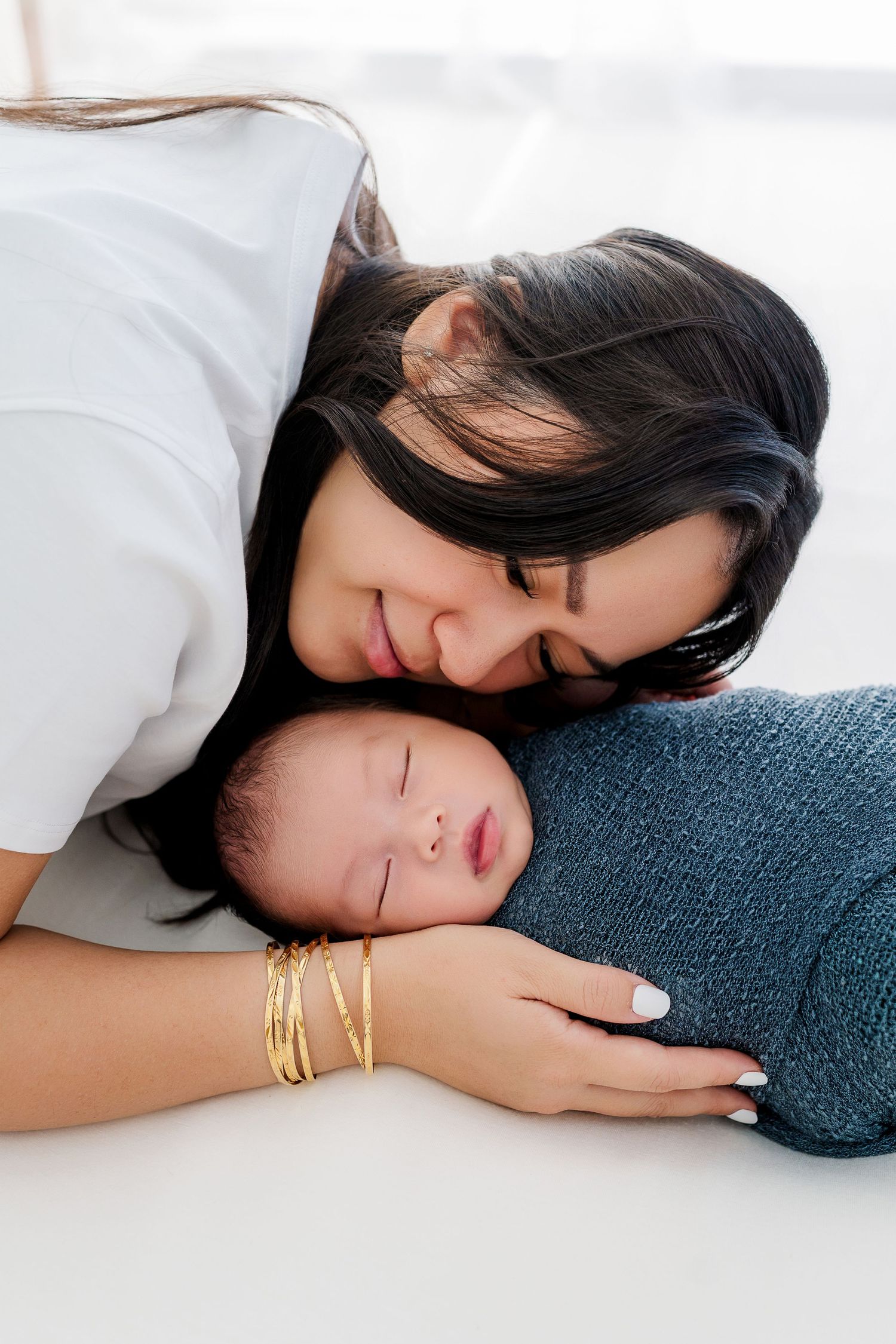 A mother with dark hair and gold bracelets tenderly rests her cheek on her sleeping newborn wrapped in a teal swaddle.